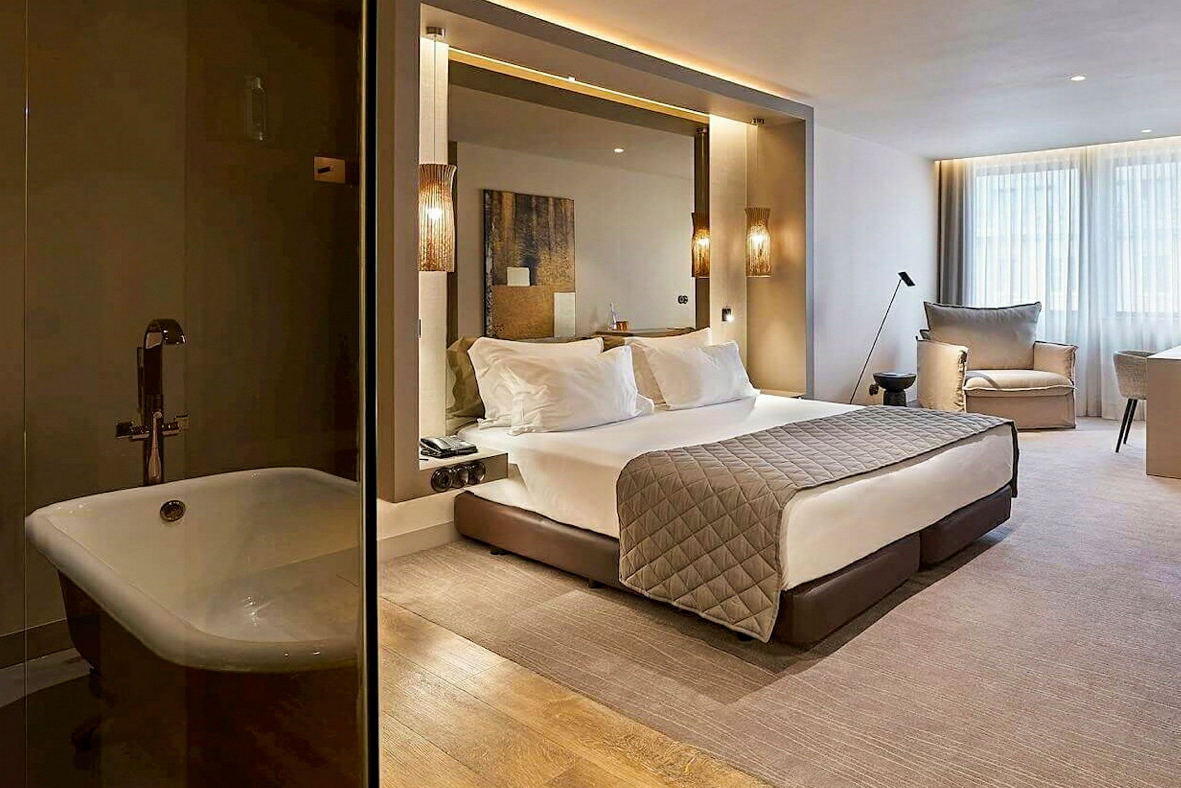 Moderni hotellihuone, jossa on suuri sänky, istuimia ja ikkunat. Vasemmalla näkyy kylpyamme. (Tekoälyn luoma teksti)