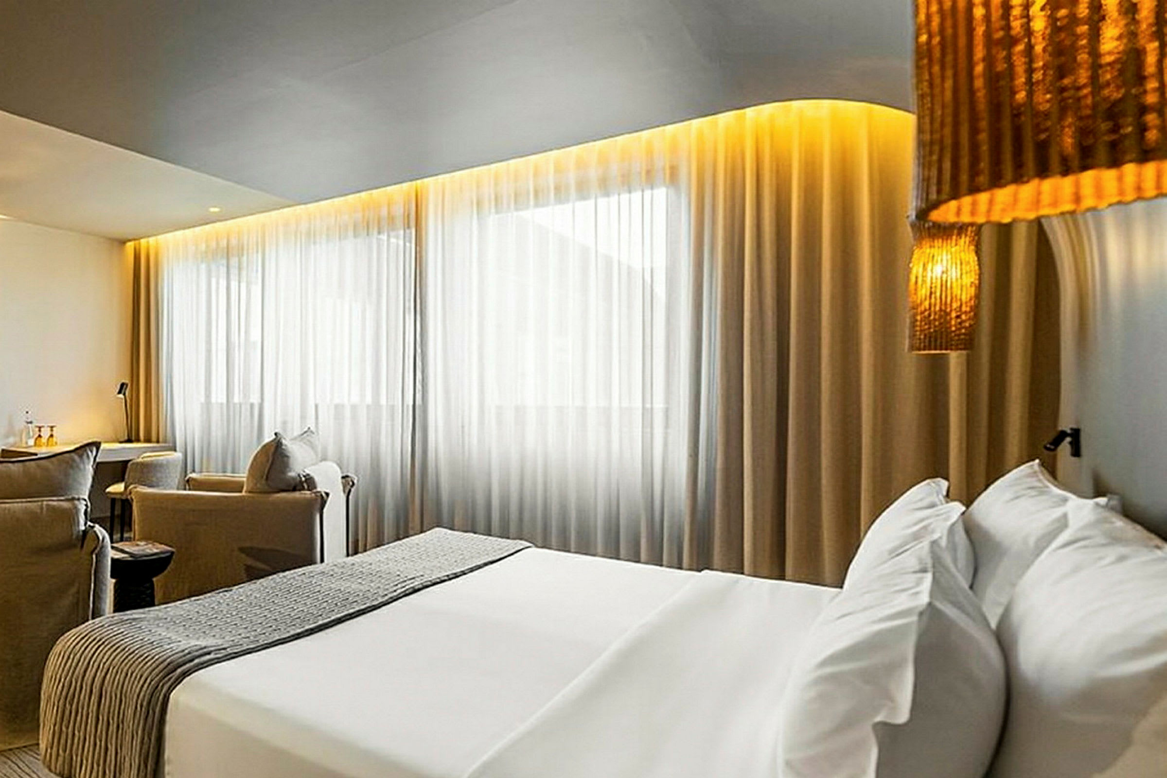 Hotellihuone, jossa on sänky, istuinryhmä ja suuret ikkunat verhoilla. (Tekoälyn luoma teksti)