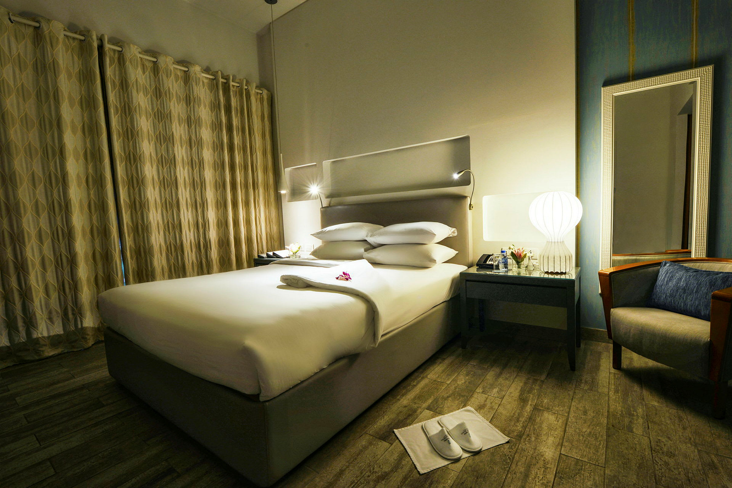 Hotellihuone, jossa on pedattu sänky, pöytä lampulla ja peilillä sekä nojatuoli vierekkäin. (Tekoälyn luoma teksti)