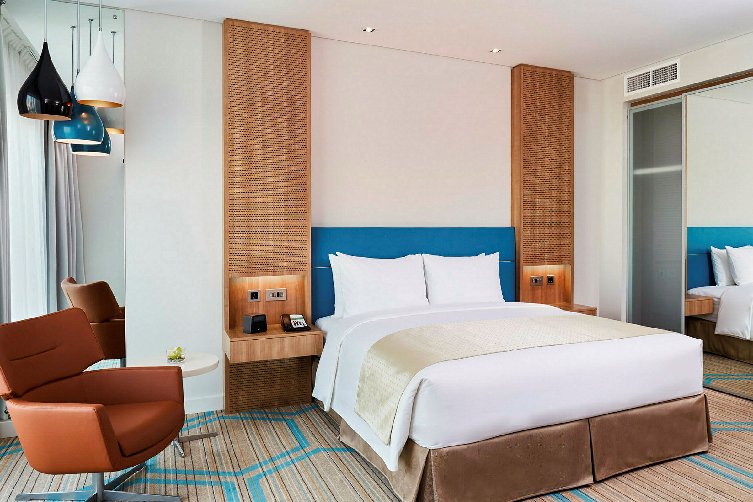 Moderni hotellihuone, jossa on parivuode, ruskea nojatuoli ja värikkäitä kattolamppuja. (Tekoälyn luoma teksti)