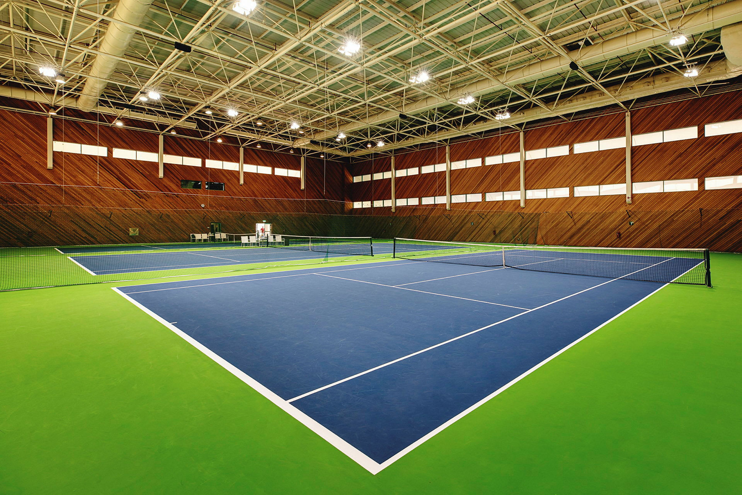 Sisätiloissa sijaitseva tenniskeskus, jossa on kaksi tenniskenttää, maalattu sinisellä ja vihreällä, ympärillä puupaneeliseinät. (Tekoälyn luoma teksti)