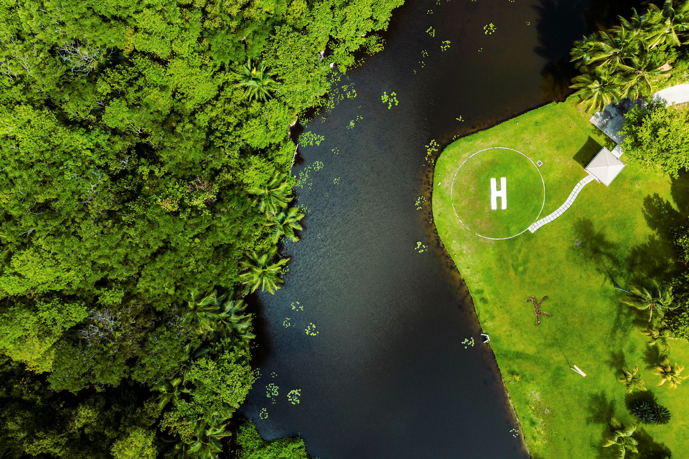 Ilmakuva vehreästä alueesta, jossa helikopterin laskeutumisalusta on merkitty "H" ja sijaitsee pienen vesialueen vieressä. (Tekoälyn luoma teksti)