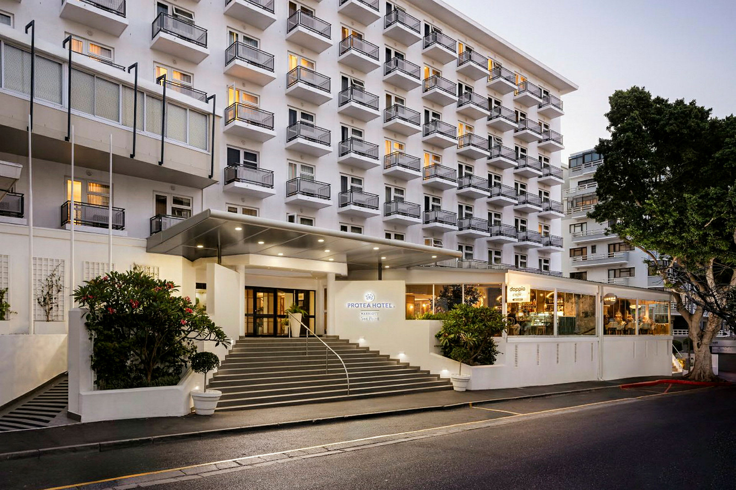 Hotellirakennus, jossa on useita kerroksia ja parvekkeita, sekä valaistu sisäänkäynti ja portaat edessä. (Tekoälyn luoma teksti)