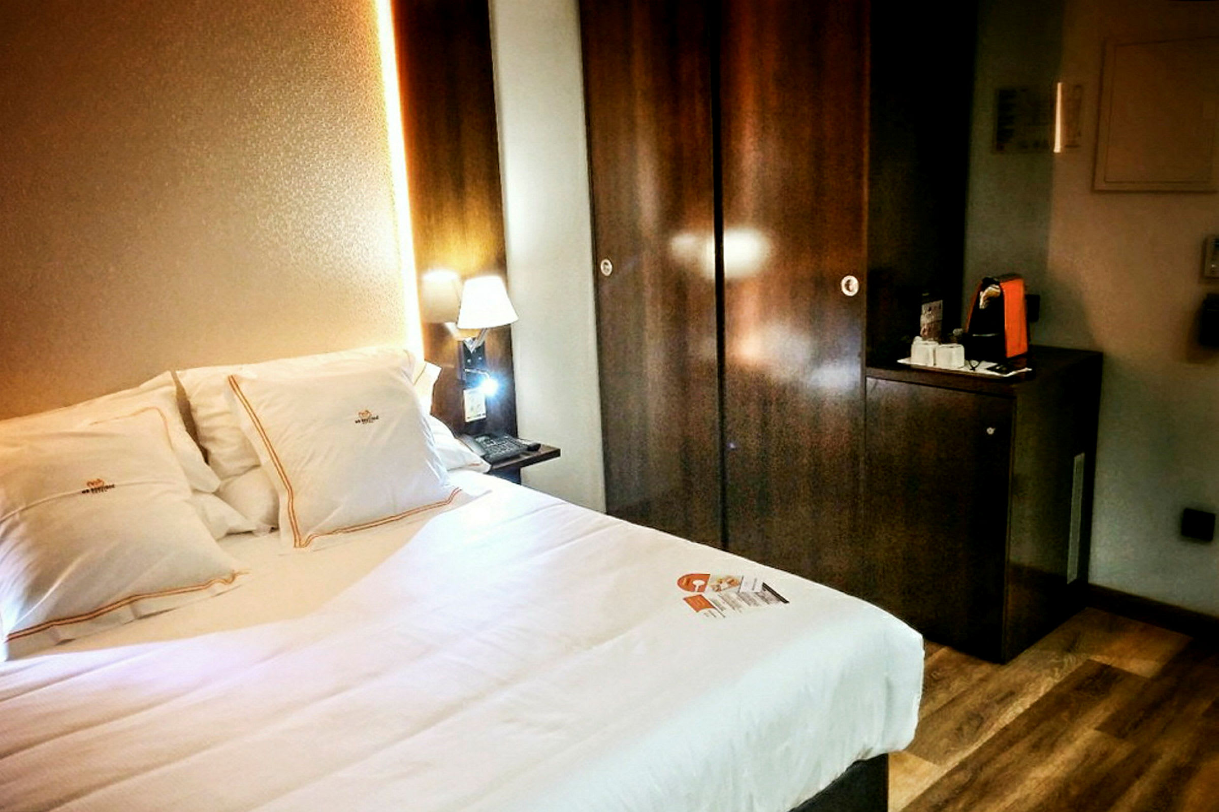 Hotellihuone, jossa sänky ja valaistu yölampu tumman puisen kaapin vieressä. (Tekoälyn luoma teksti)