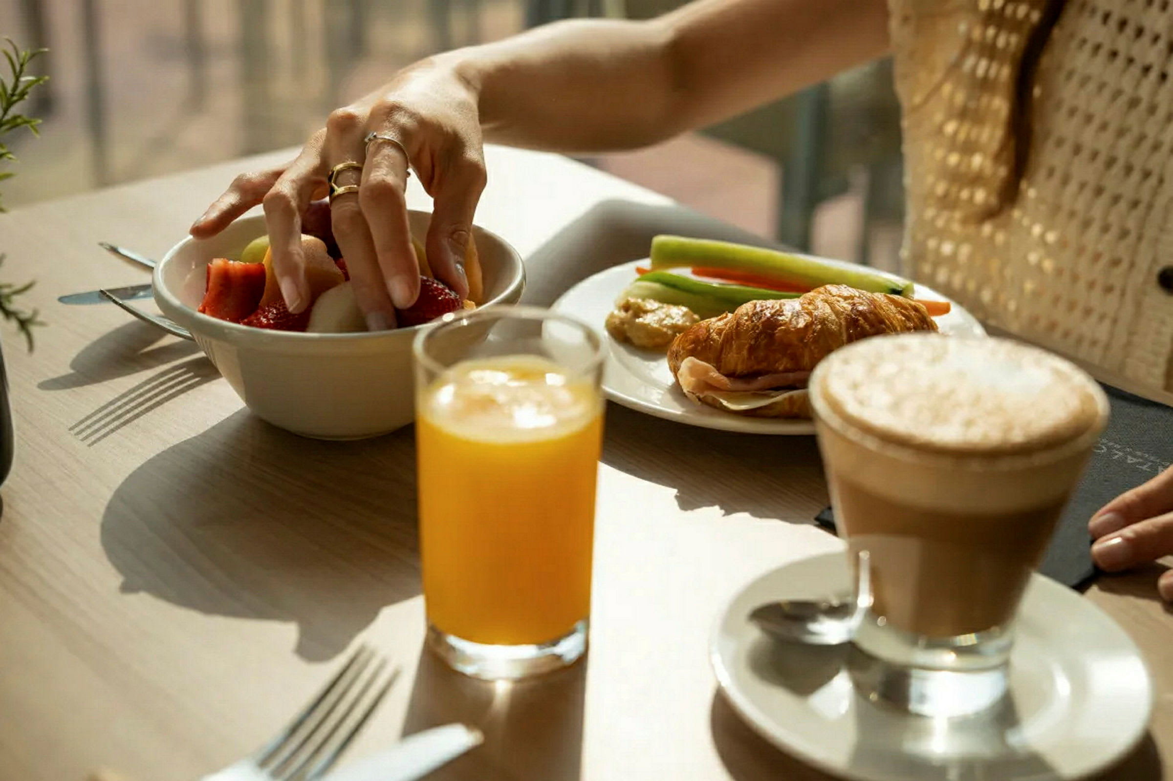 Henkilö ottaa hedelmää kulhosta pöydällä, jolla on croissant, vihanneksia, mehua ja kahvia. (Tekoälyn luoma teksti)