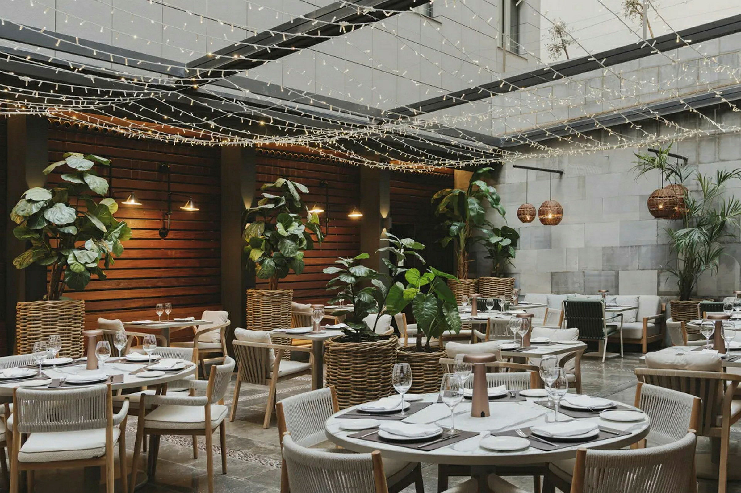 Ravintola ulkoalueella, pöydät koristeltu aterioita varten, ympäröitynä kasveilla ja valonauhoilla. (Tekoälyn luoma teksti)