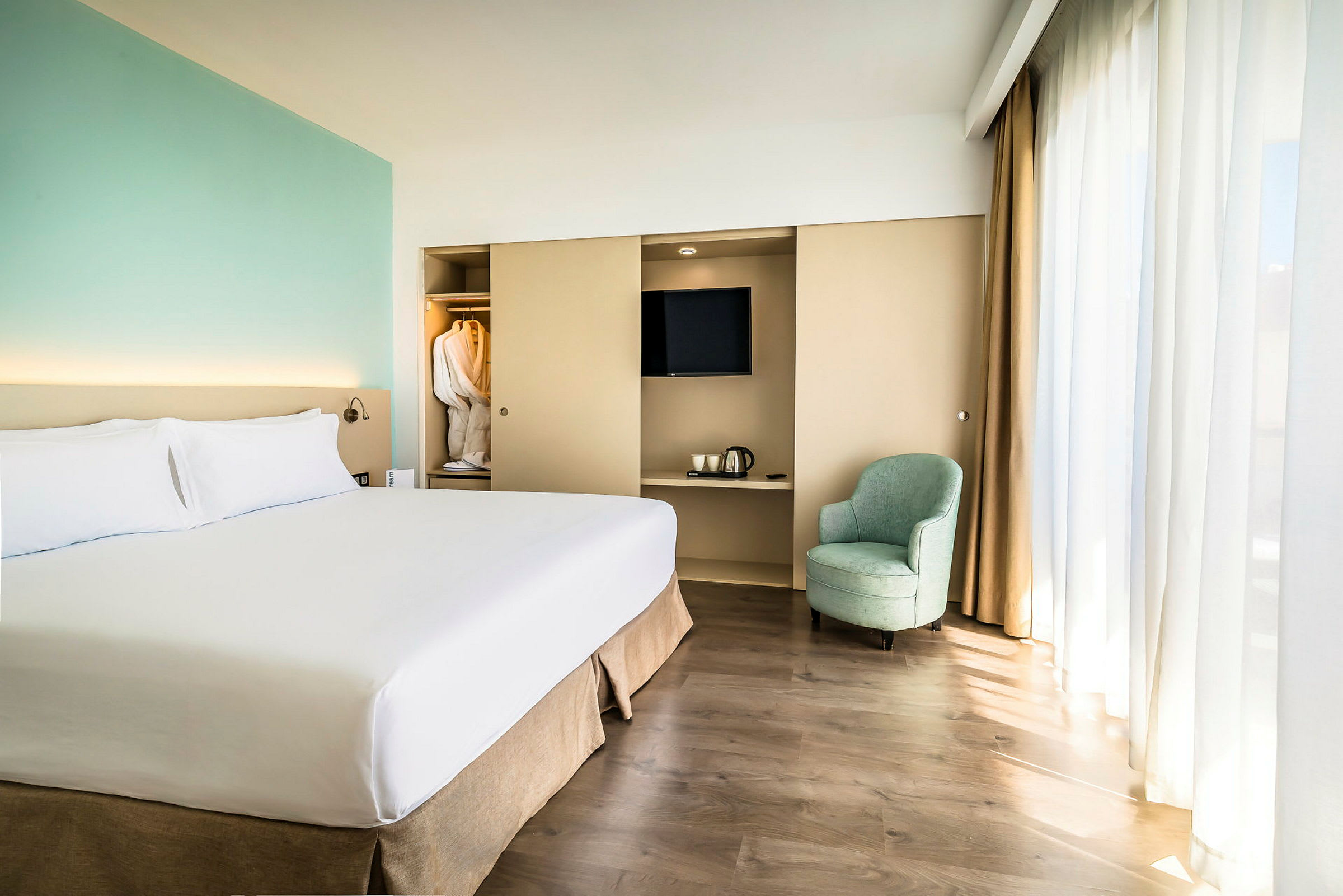 Hotellihuone, jossa on suuri sänky, nojatuoli, kaappi, jossa televisio ja vedenkeitin, sekä vaatekaappi, jossa on kylpytakkeja. (Tekoälyn luoma teksti)