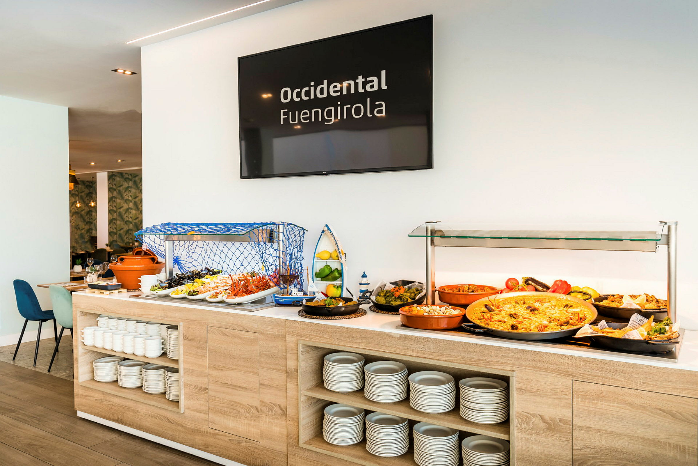 Buffet, jossa on erilaisia ruokia ja lautasia, sekä seinällä kyltti, jossa lukee 'Occidental Fuengirola'. (Tekoälyn luoma teksti)