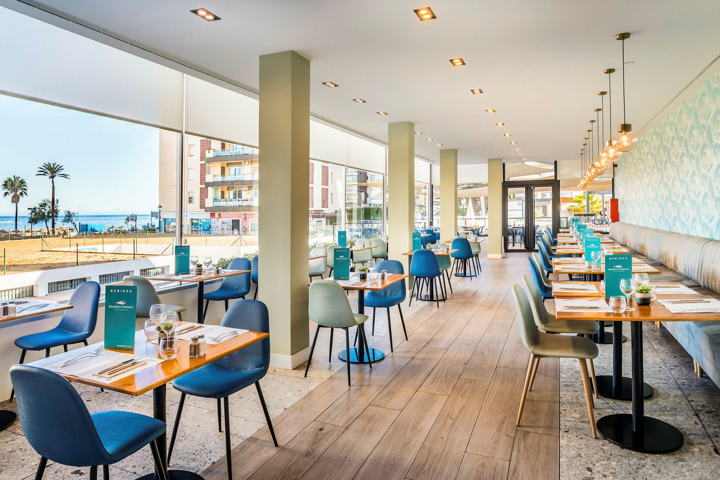 Raikas ja moderni ravintola, jossa on suuret ikkunat, jotka tarjoavat näkymän merelle. Pöydät on katettu, ja tuolit ovat erivärisiä. (Tekoälyn luoma teksti)