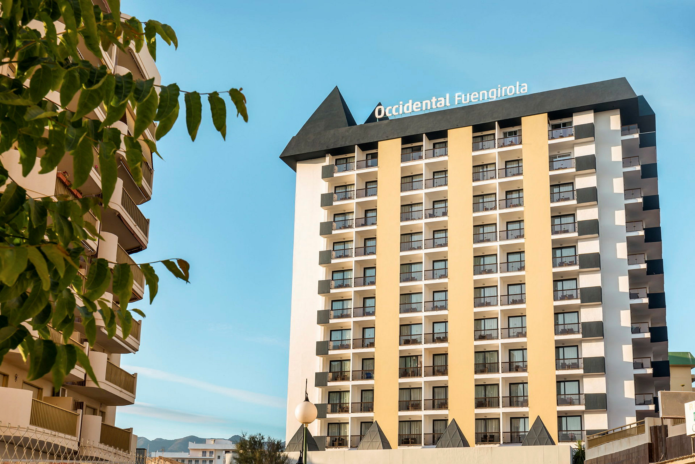 Kuva hotellista, jossa on monta kerrosta ja parveketta, ympärillä puita ja kirkas sininen taivas. (Tekoälyn luoma teksti)