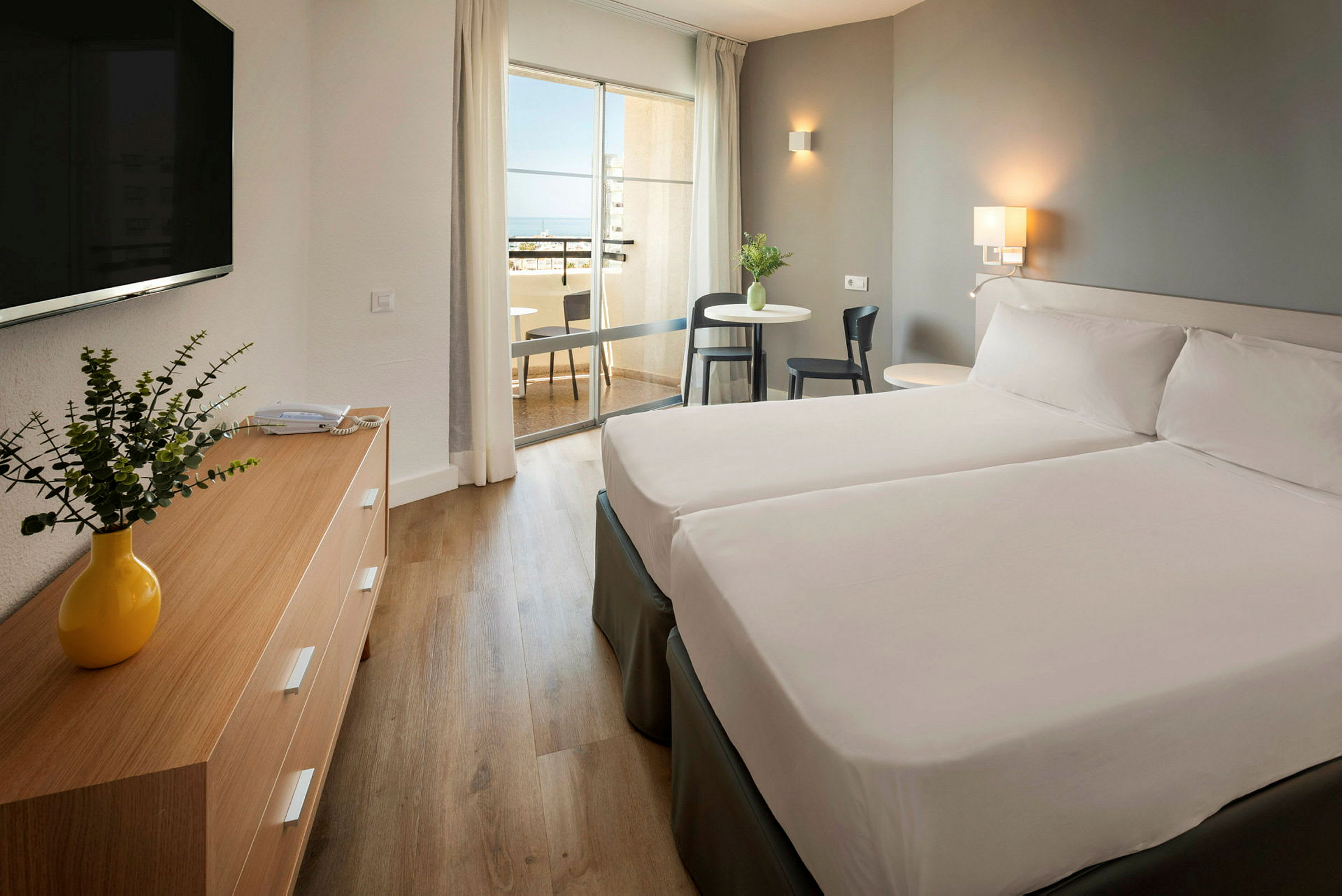 Hotellihuone, jossa on kaksi sänkyä, puinen pöytä keltaisella maljakolla ja parveke taustalla. (Tekoälyn luoma teksti)