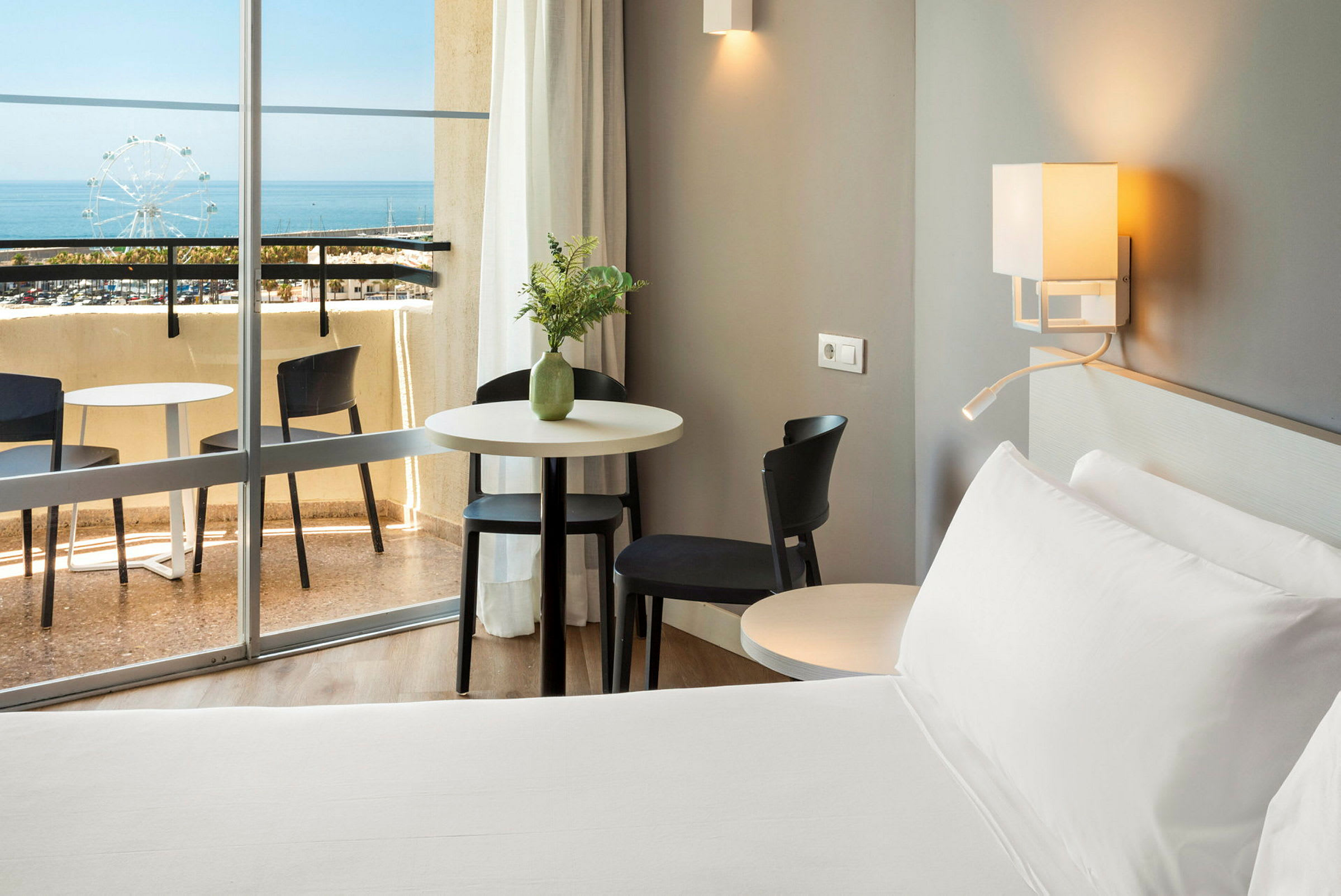 Hotellihuone, jossa vuode, pöytä ja tuolit lähellä parveketta merinäköalalla. (Tekoälyn luoma teksti)