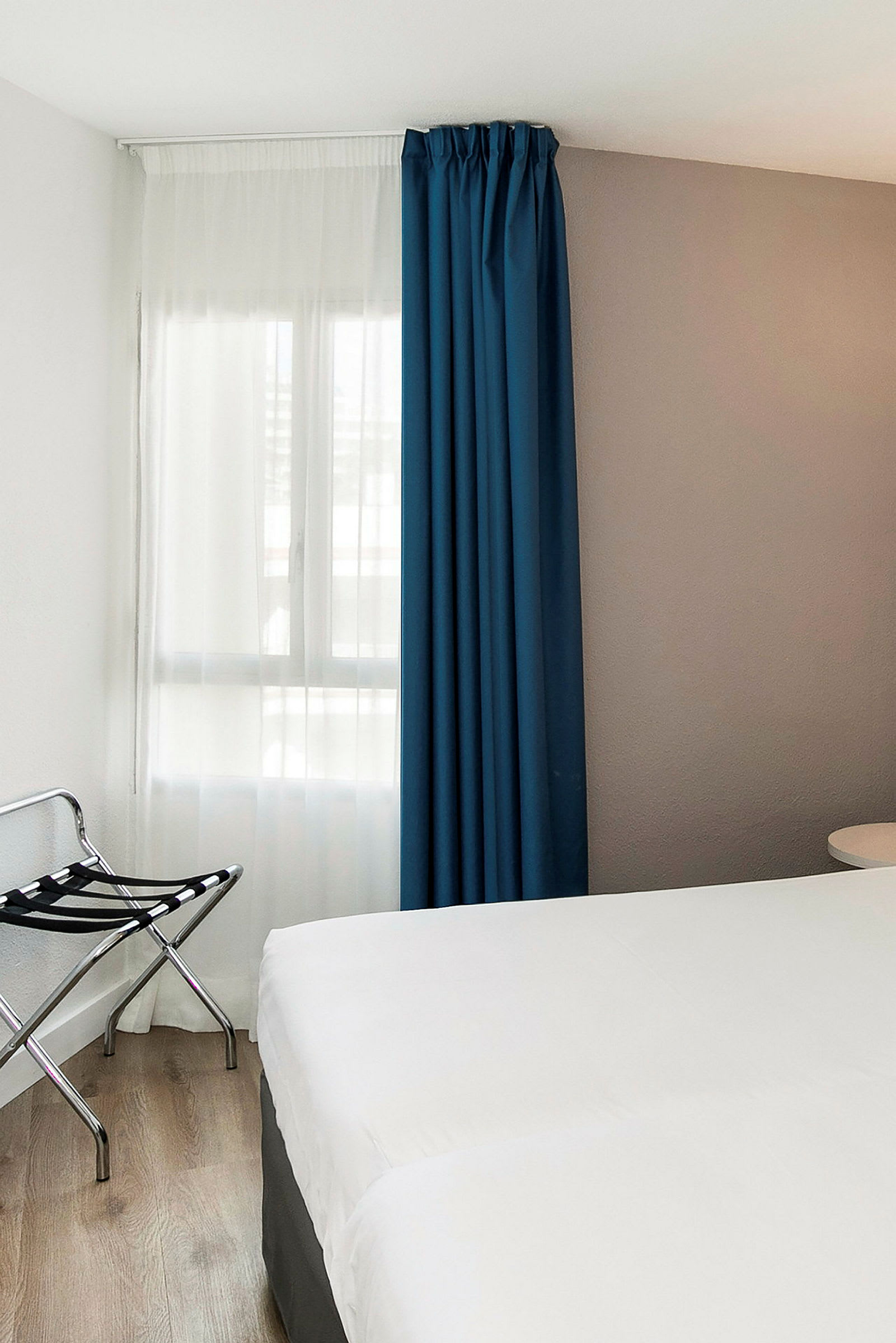 Hotellihuone, jossa valkoinen sängynpeite, sininen verho ja matkatavarahylly vieressä. (Tekoälyn luoma teksti)