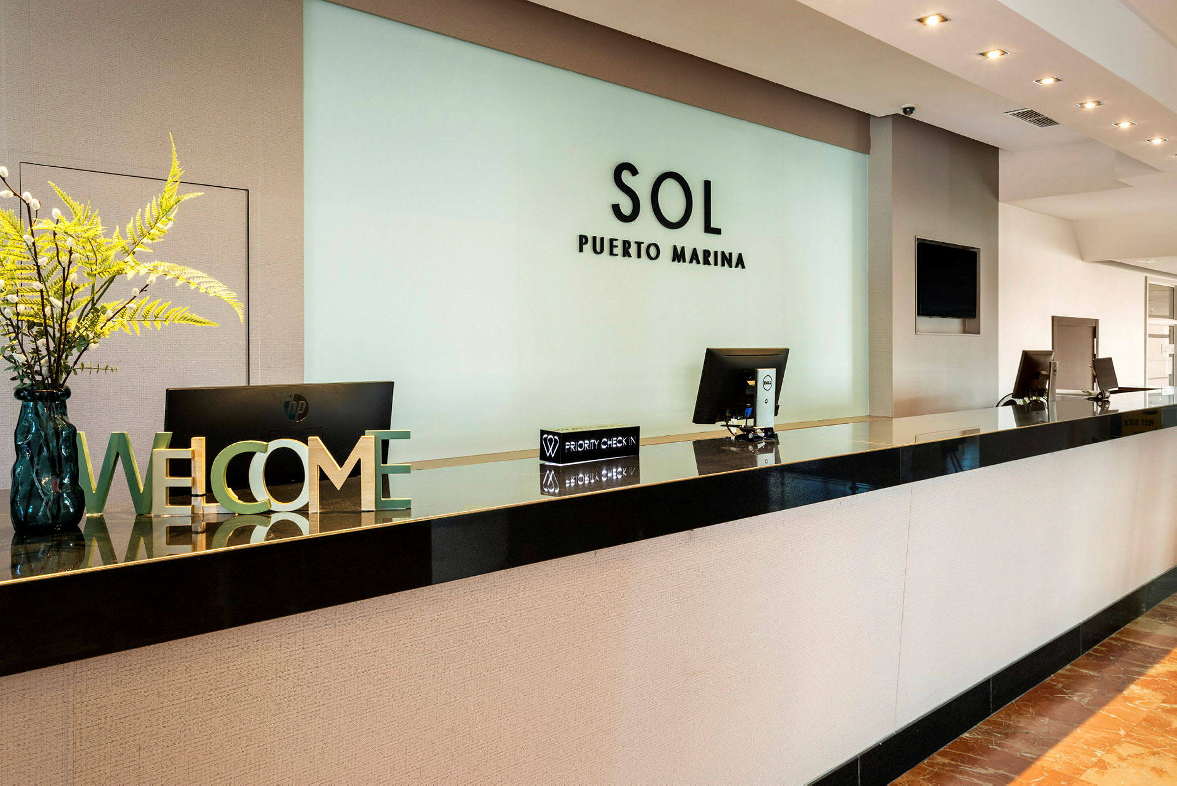 Sol Puerto Marina -hotellin vastaanotto, jossa on tietokoneen näytöt ja koristeet, mukaan lukien tervetuliaiskyltti ja kukka-asetelma. (Tekoälyn luoma teksti)