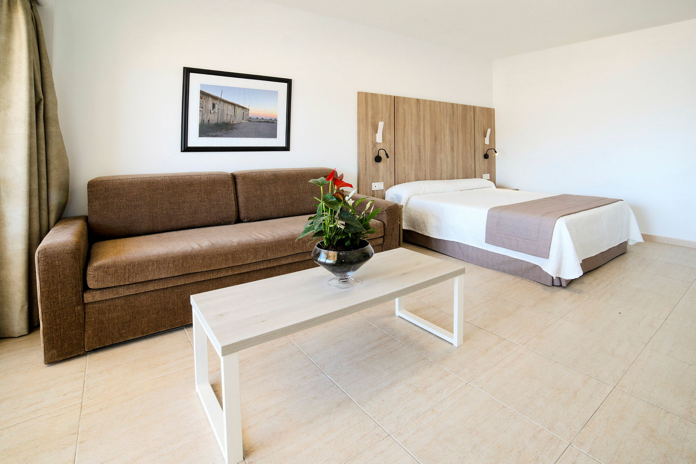 Hotellihuone, jossa ruskea sohva, pöytä kukkamaljakolla ja sänky, jonka takana on puupaneeli. (Tekoälyn luoma teksti)