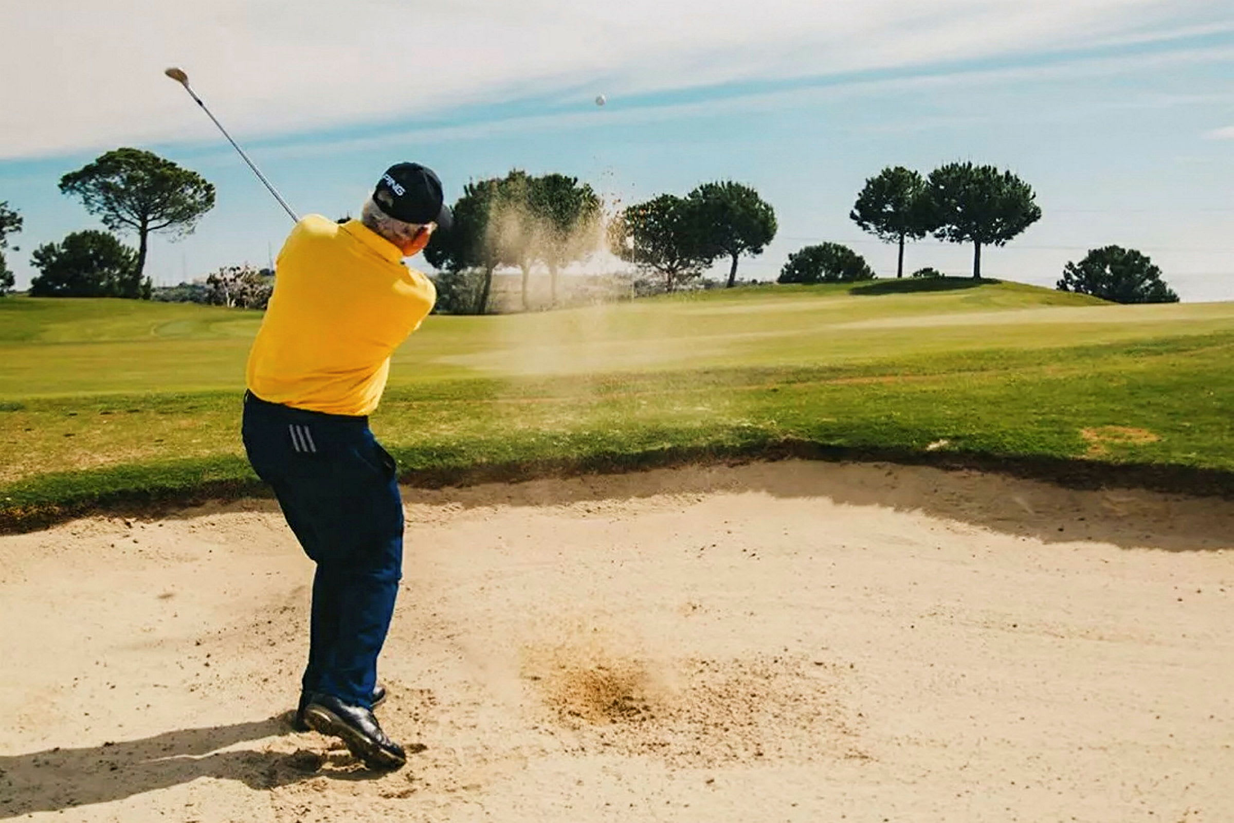 Henkilö lyö golfpalloa hiekkaesteestä golfkentällä, jota ympäröivät vihreät puut. (Tekoälyn luoma teksti)