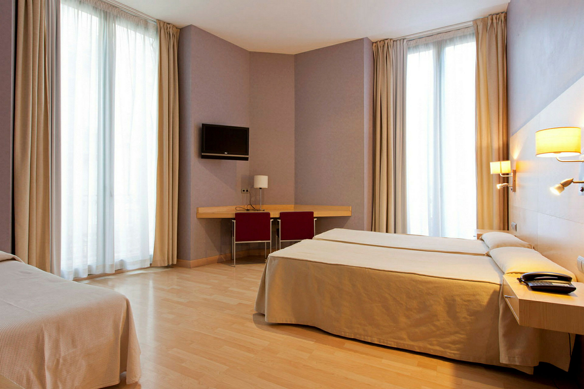 Hotellihuone, jossa on kaksi vuodetta, kirjoituspöytä ja tuoli, seinään kiinnitetty televisio sekä suuret ikkunat verhoilla. (Tekoälyn luoma teksti)