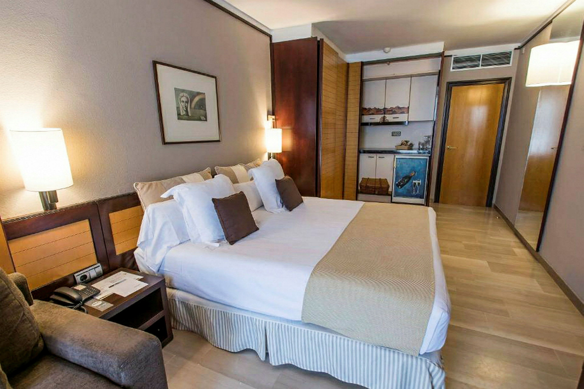 Hotellihuone, jossa on iso sänky, tyynyjä, yöpöytä ja pieni keittiöosa kaappeineen. (Tekoälyn luoma teksti)