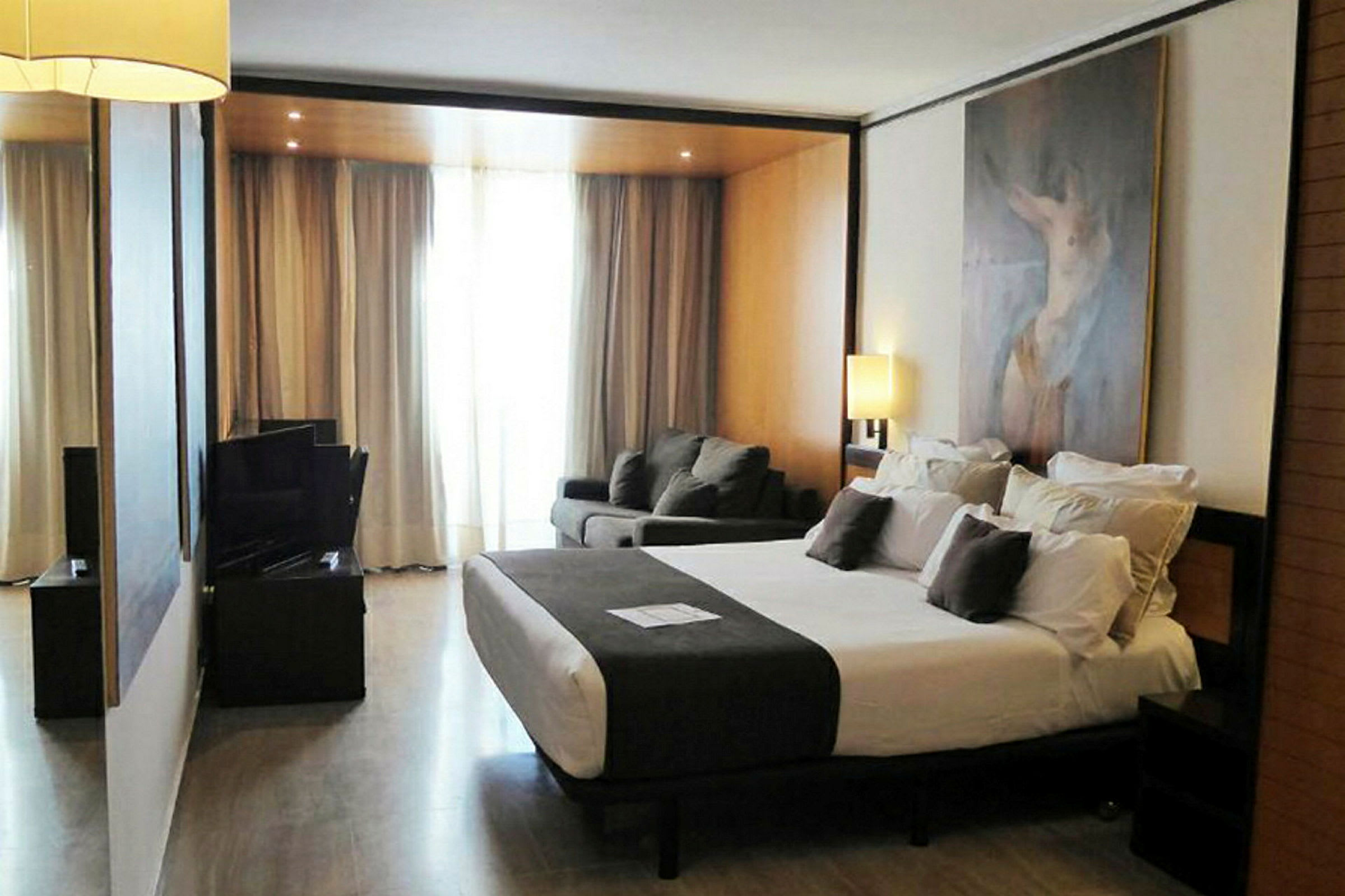 Hotellihuone, jossa suuri sänky, istuinryhmä ja lamppuja sekä seinällä maalaus. (Tekoälyn luoma teksti)