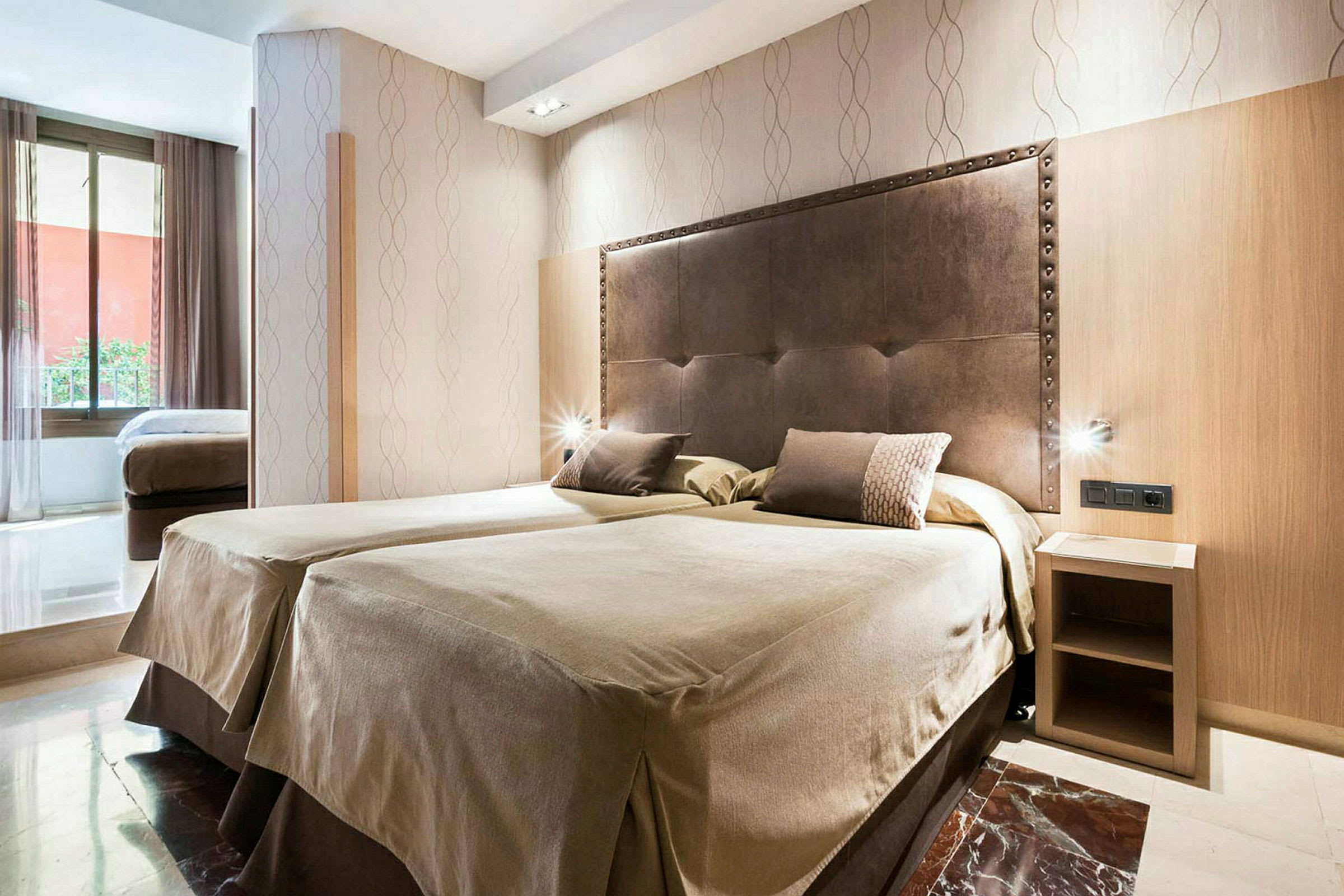 Hotellihuone, jossa on kaksi vierekkäistä yhden hengen vuodetta ja tummaa materiaalia oleva sängynpääty. Huoneessa on pieni yöpöytä ja suuri ikkuna, josta tulee luonnonvaloa. (Tekoälyn luoma teksti)