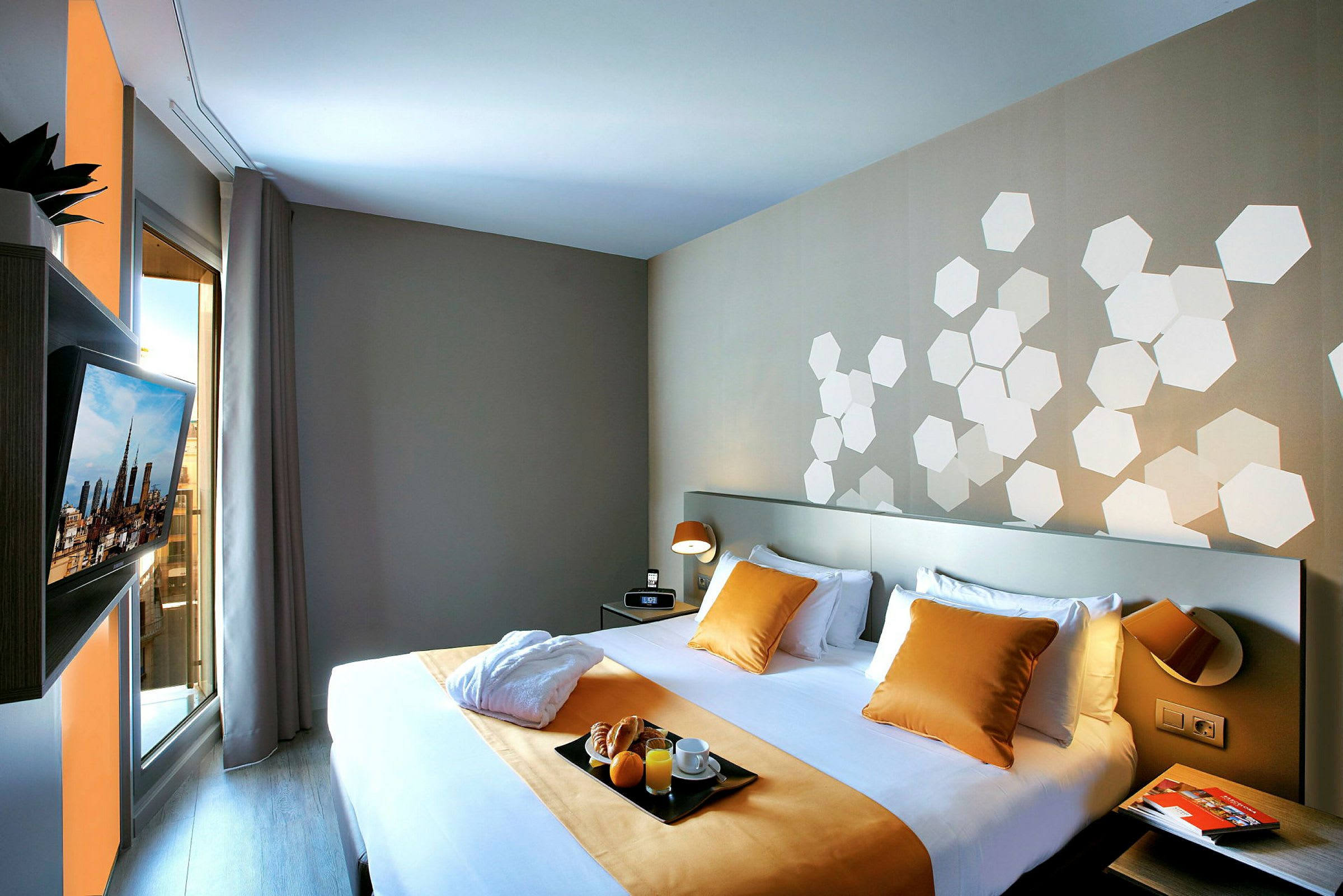 Hotellihuone, jossa on parisänky, vuodevaatteina valkoiset lakanat sekä keltaiset tyynyt ja peitto. Seinällä on valkoisia kuusikulmiokuvioita. Näkyvissä on myös ikkuna verhoineen sekä seinälle kiinnitetty televisio. (Tekoälyn luoma teksti)