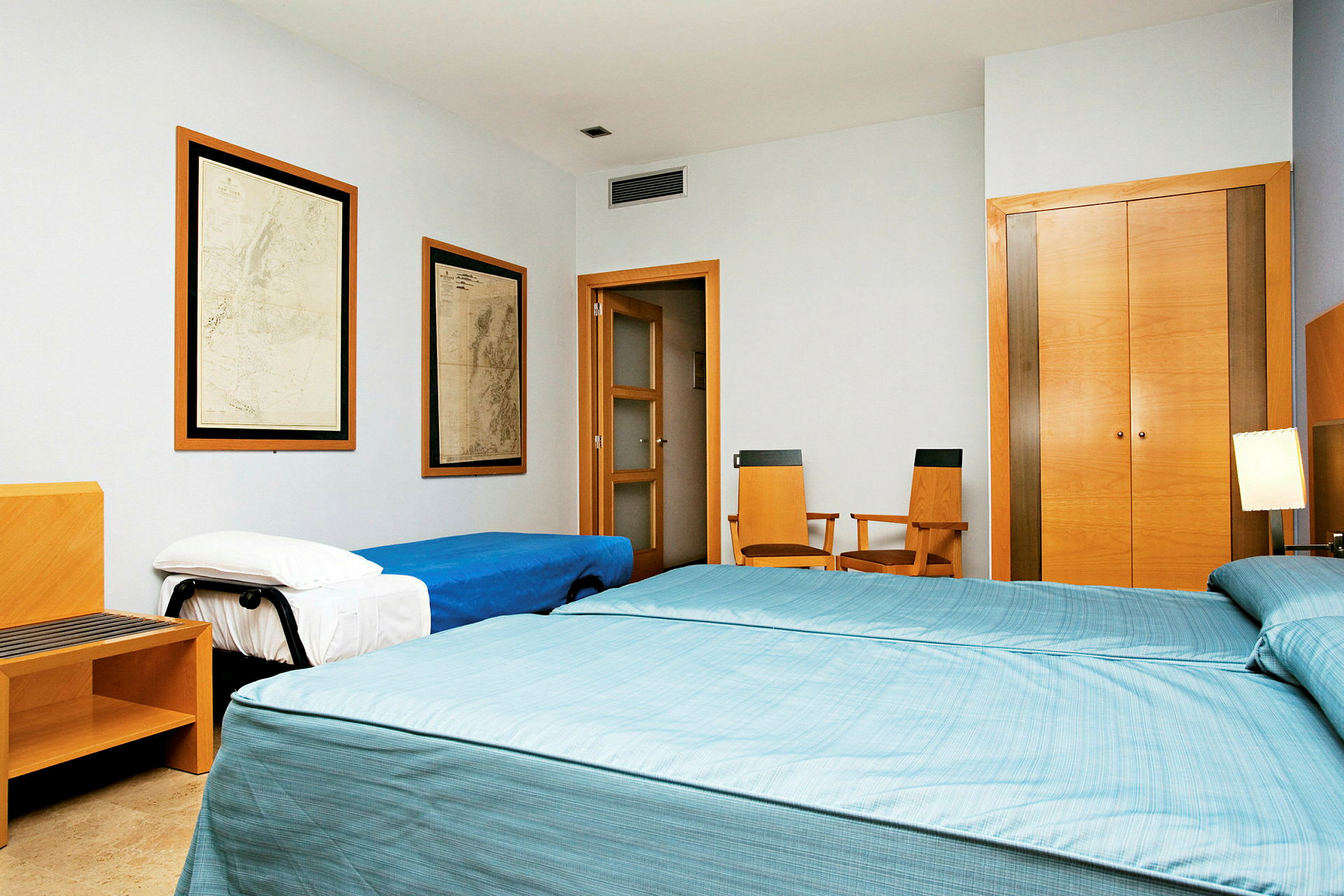 Makuuhuone, jossa on kaksi yhden hengen vuodetta sinisillä päiväpeitteillä, kolmas sänky sinisellä päiväpeitteellä sivussa, kaksi puutuolia, kaappi kaksinkertaisilla puuovilla ja kaksi taulua seinällä. (Tekoälyn luoma teksti)