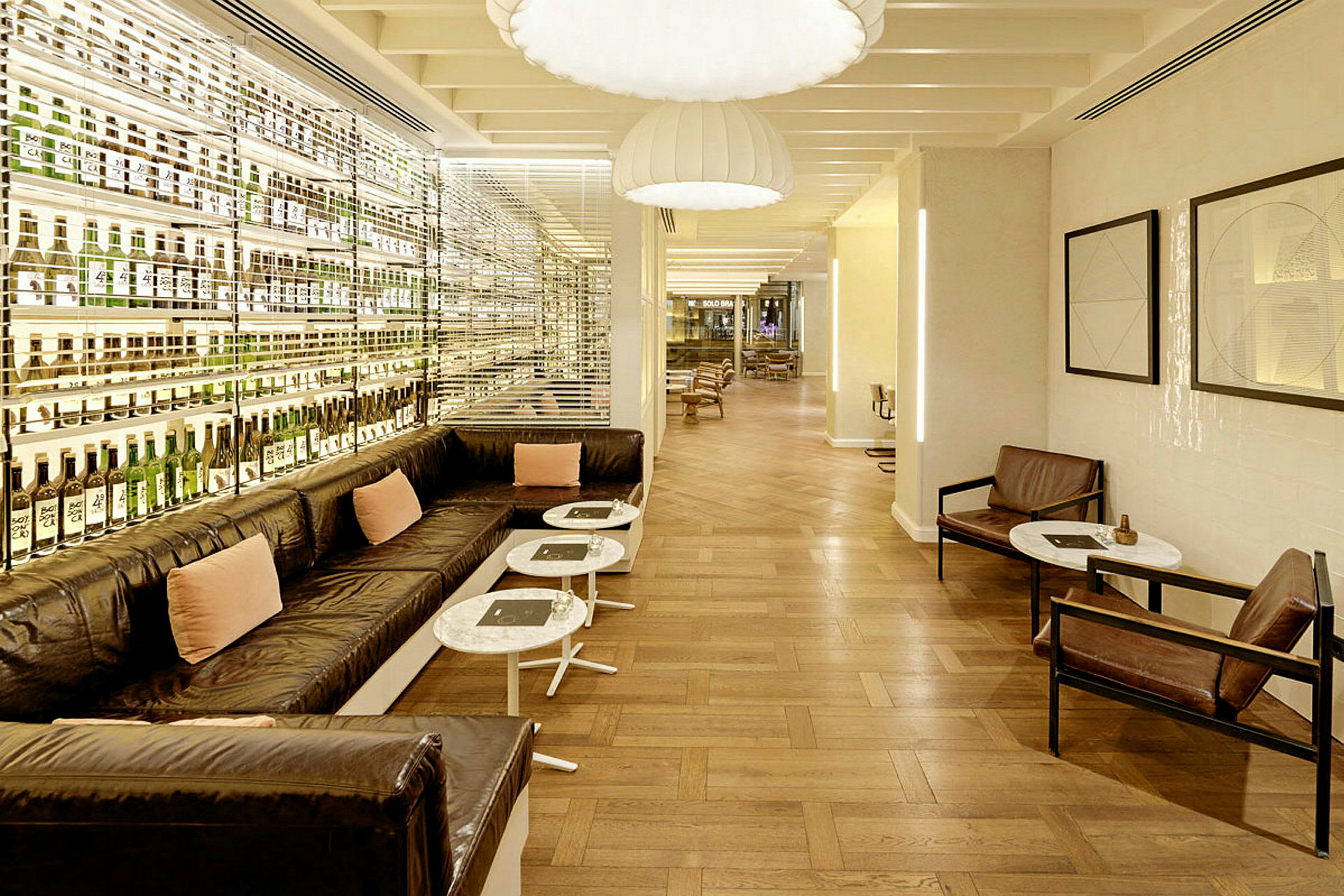 Moderni sisustettu lounge-alue, jossa on pitkä sohva, tuoleja, pieniä pöytiä ja pulloja seinähyllyillä. (Tekoälyn luoma teksti)