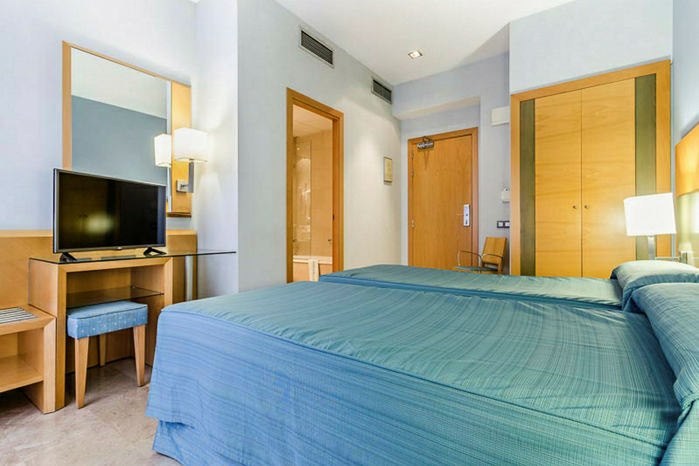 Hotellihuone, jossa on vuode sinisellä päiväpeitteellä, pöytä peilillä ja televisio päällä sekä vaatekaapit, kylpyhuoneen ovi ja ulko-ovi. (Tekoälyn luoma teksti)