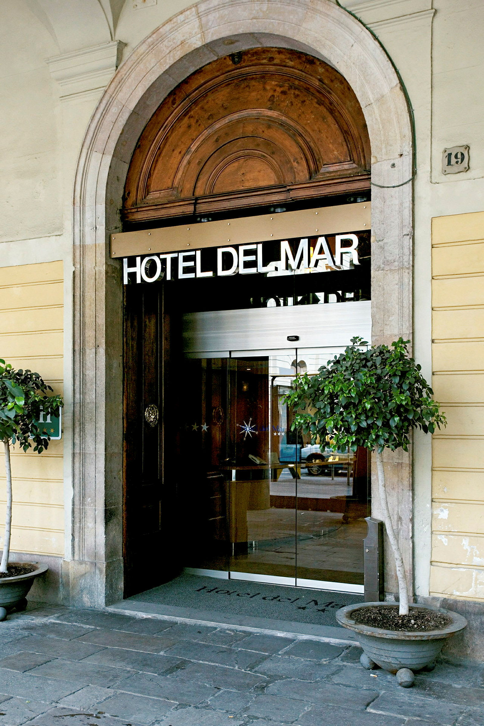 Sisäänkäynti hotelliin, jossa lukee 'HOTEL DEL MAR'. Sisäänkäynti on kaareva lasiovilla, ja kummallakin puolella on koristeellisia kukkaruukkuja. (Tekoälyn luoma teksti)