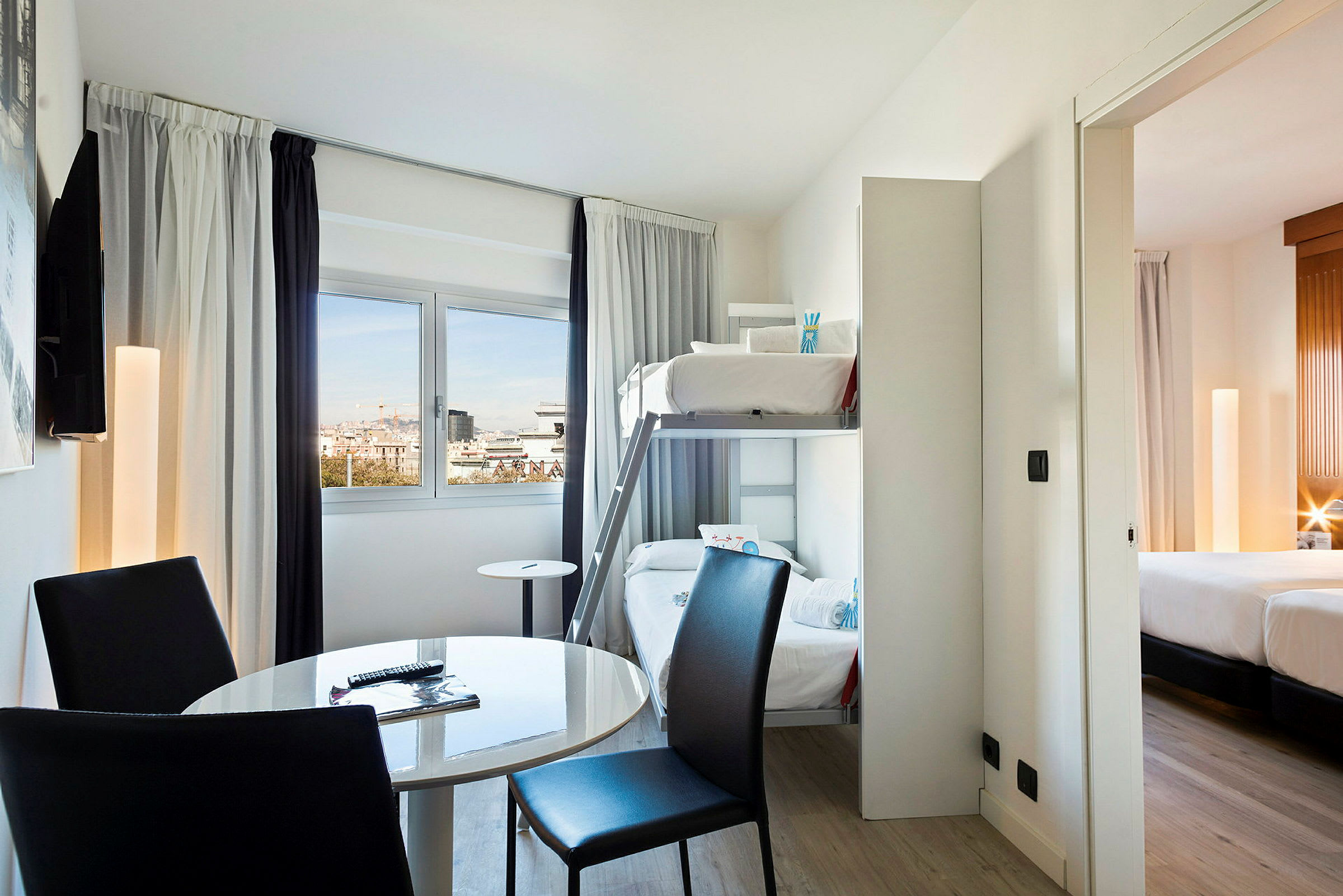Hotellihuone, jossa on kerrossängyt, pyöreä pöytä ja tuolit sekä näkymä ikkunan läpi. Viereinen makuuhuone, jossa parisänky, näkyy. (Tekoälyn luoma teksti)