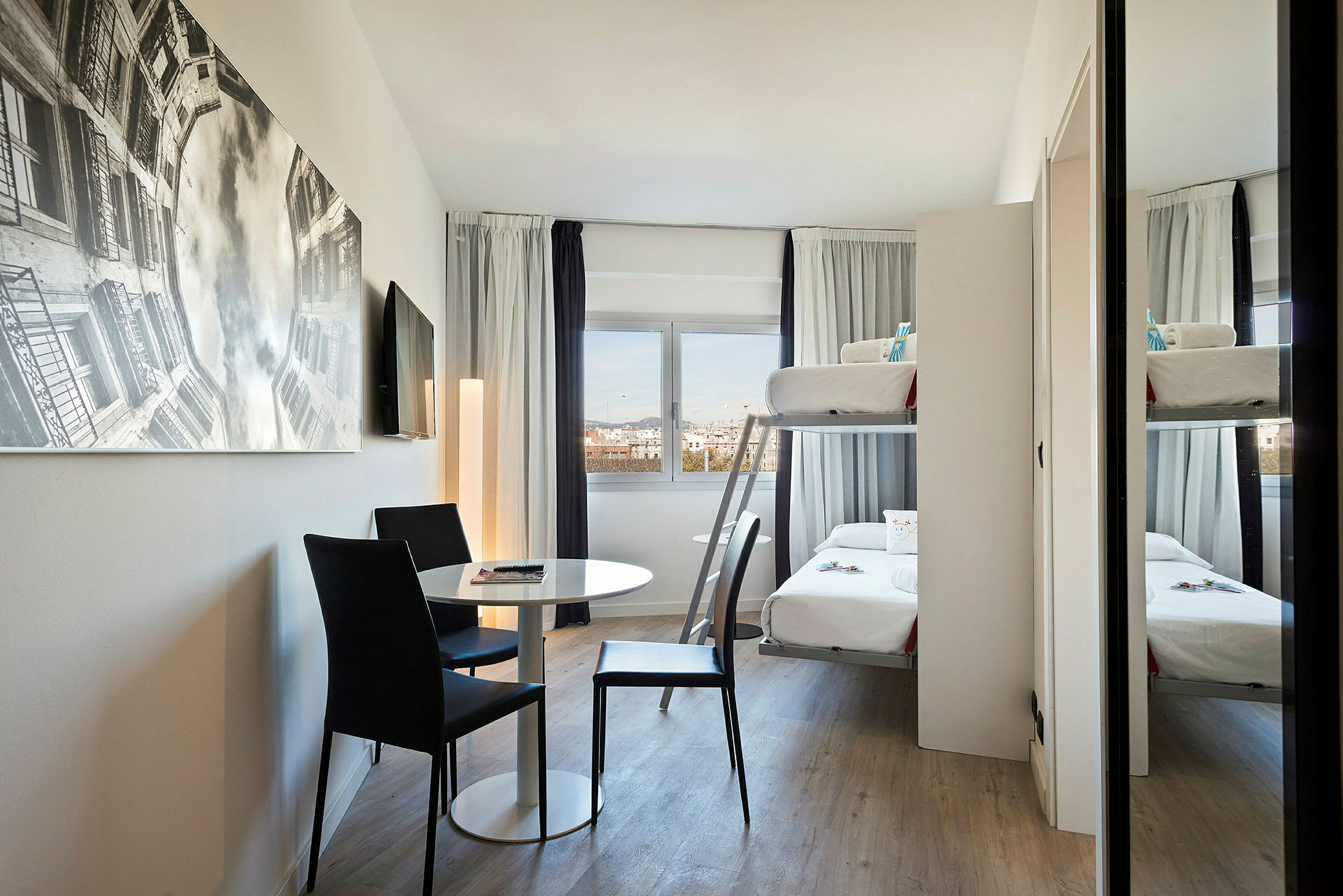 Moderni hotellihuone, jossa on kerrossänky, pöytä ja kaksi tuolia sekä suuri seinäkuva. (Tekoälyn luoma teksti)