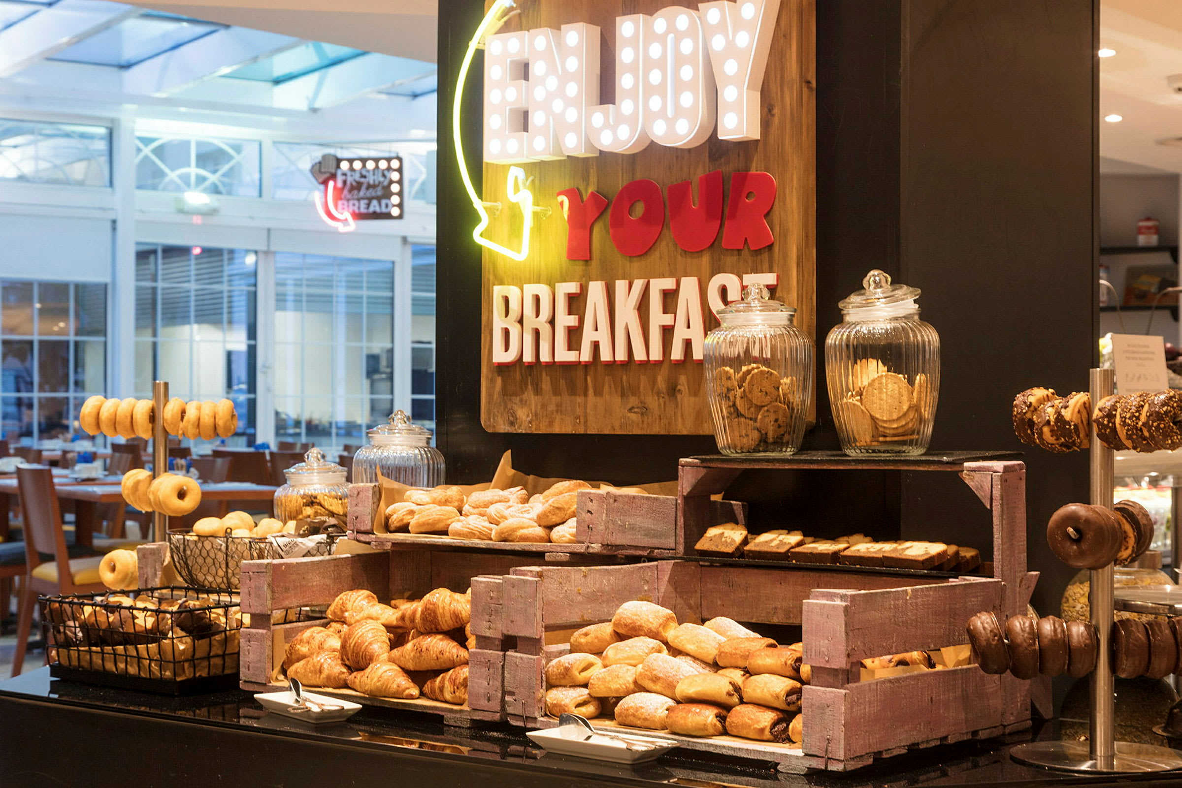 Buffet, jossa on erilaisia aamiaisherkkuja, kuten croissanteja, donitseja ja pikkuleipiä, koristeltuna kylteillä, jotka kannustavat nauttimaan aamiaisesta. (Tekoälyn luoma teksti)