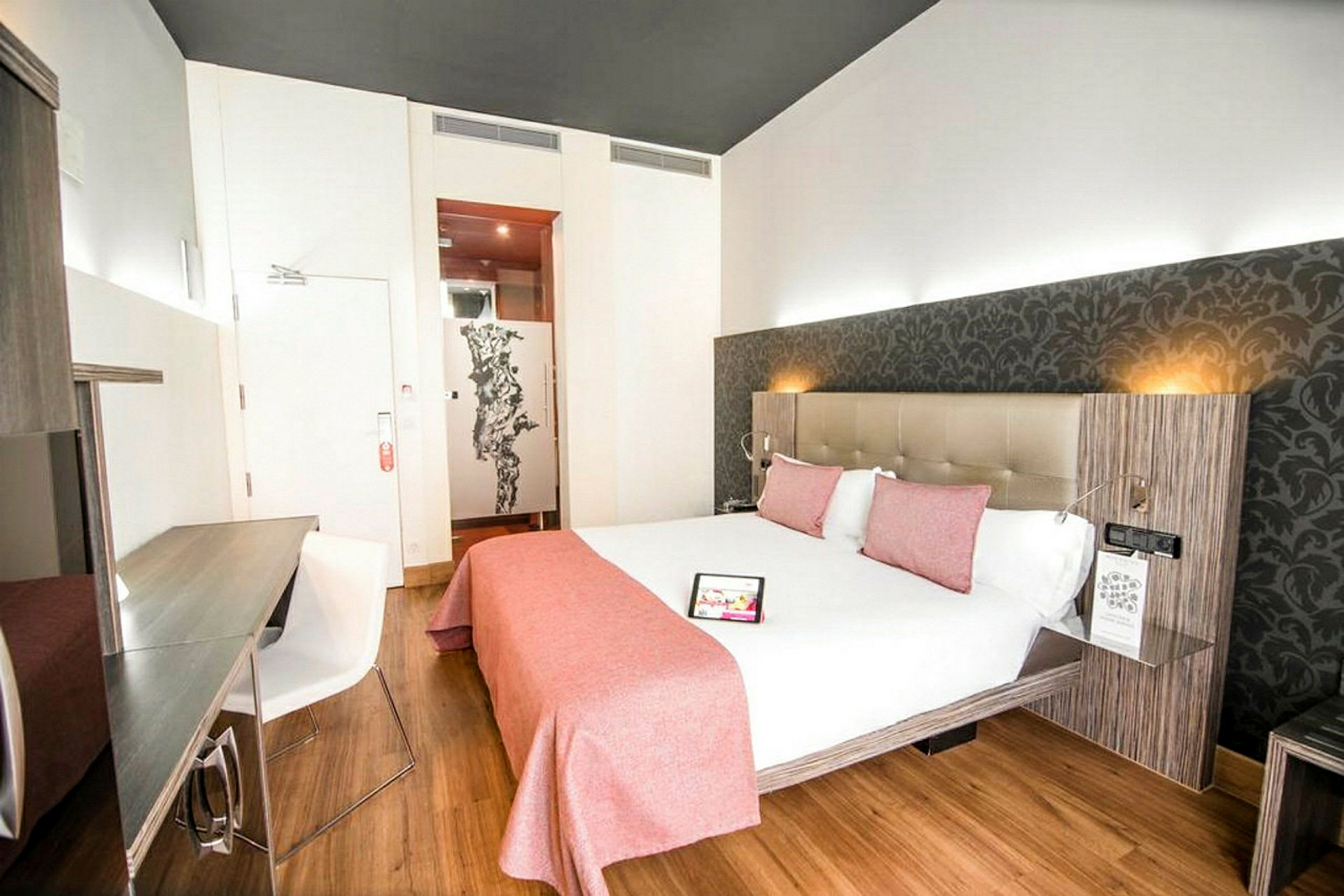 Valoisa hotellihuone, jossa on parisänky valkoisilla lakanoilla ja vaaleanpunaisilla vuodevaatteilla, kirjoituspöytä tuoleineen ja koristeellinen seinä sängyn takana. (Tekoälyn luoma teksti)