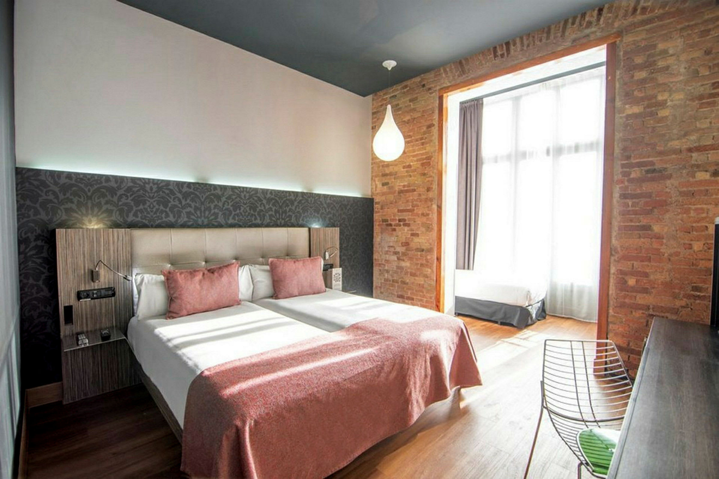 Hotellihuone, jossa on kaksi erillistä vuodetta, vaaleanpunaiset päiväpeitot ja tiiliseinä suuren ikkunan vieressä. (Tekoälyn luoma teksti)
