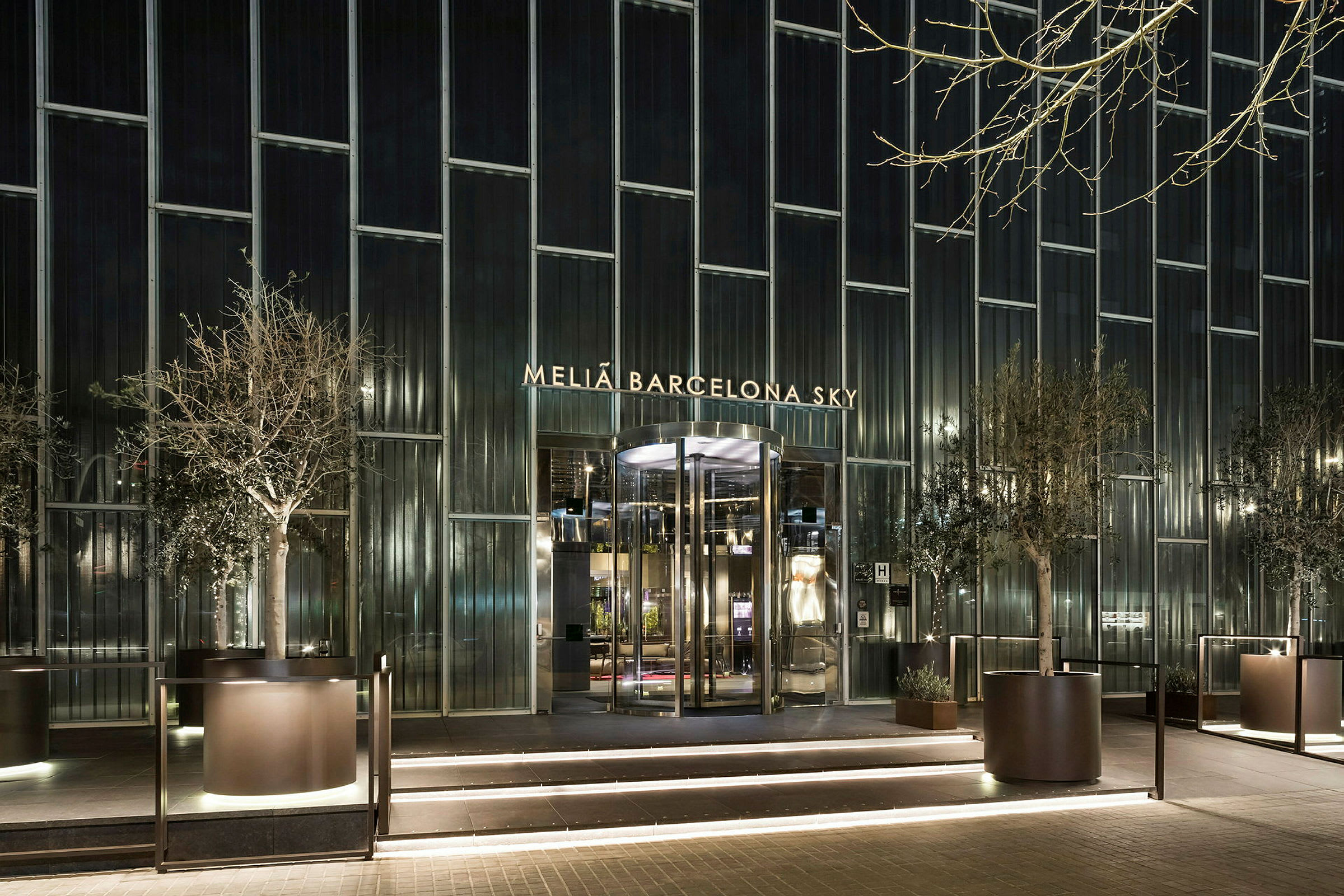 Melia Barcelona Sky -hotellin julkisivu, jossa valaistu sisäänkäynti ja suuret lasi-ikkunat, ympäröitynä istutetuilla puilla. (Tekoälyn luoma teksti)