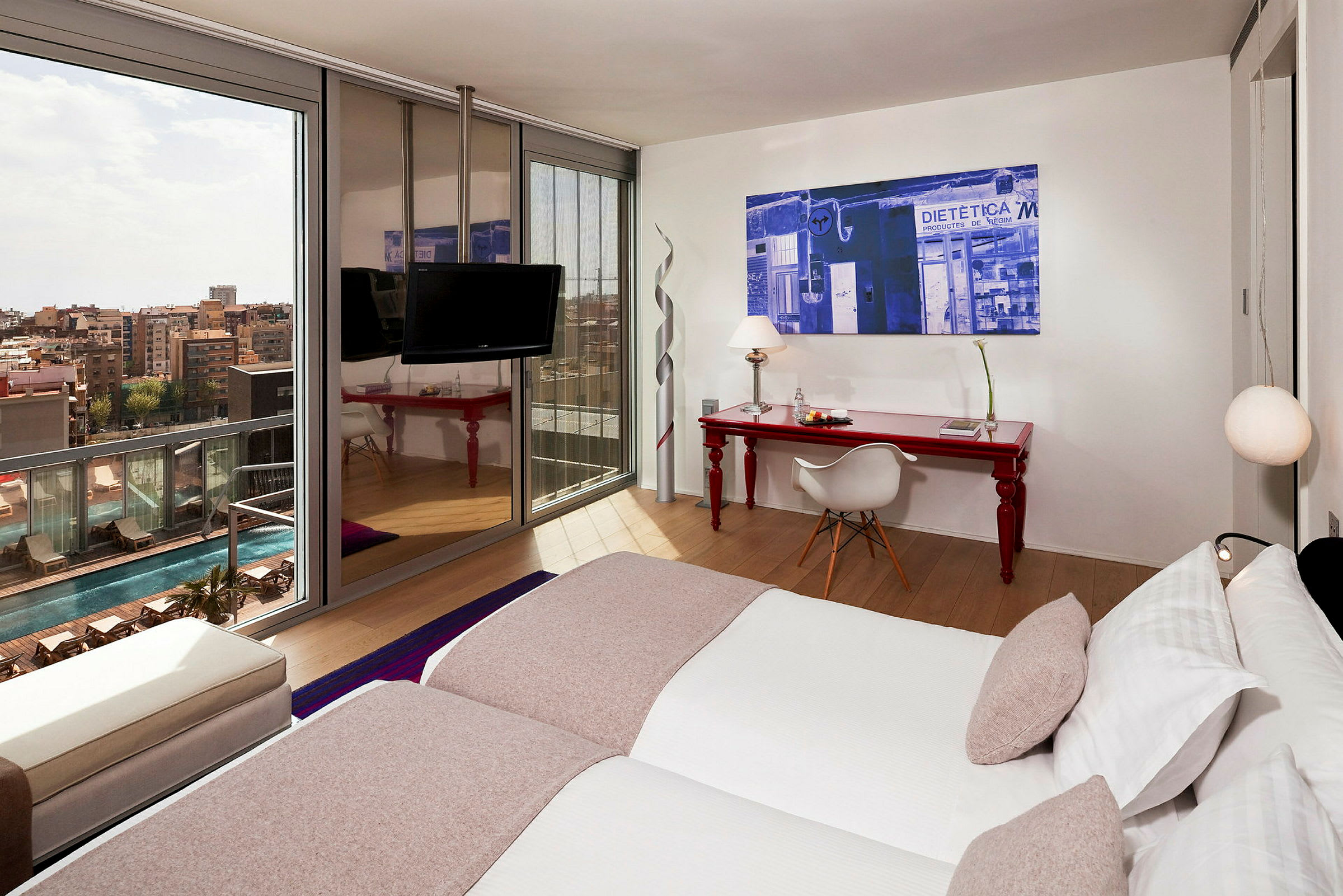 Moderni hotellihuone, jossa on kaksi erillistä vuodetta, punainen työpöytä tuolilla ja suuri ikkuna, josta avautuu näkymä uima-allasalueelle ja kaupunkiin. (Tekoälyn luoma teksti)
