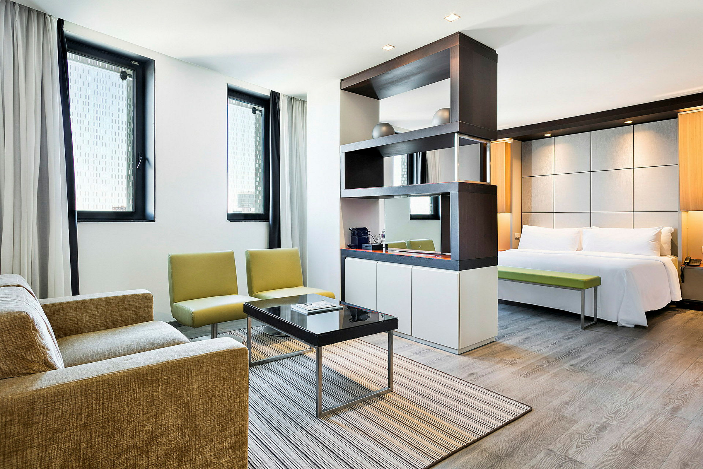 Moderni hotellihuone, jossa on olohuone ja makuuhuone. Olohuoneessa on sohva, nojatuolit ja sohvapöytä. Makuuhuoneessa on parivuode ja penkki. (Tekoälyn luoma teksti)