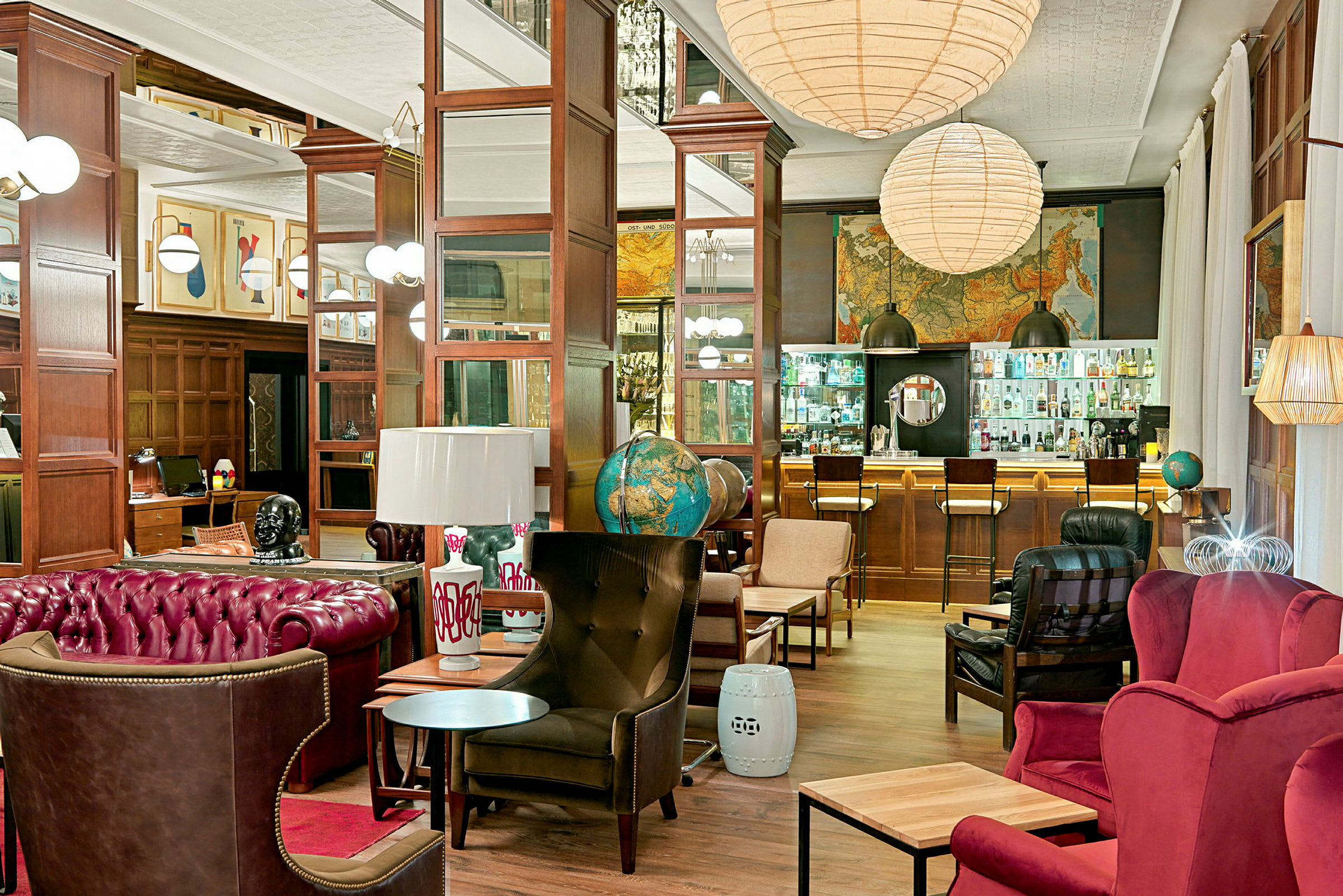 Tyylikäs lounge, jossa punaisia ja ruskeita huonekaluja, maapalloja, taideteoksia seinillä ja taustalla baari lasipintaisella tiskillä. (Tekoälyn luoma teksti)