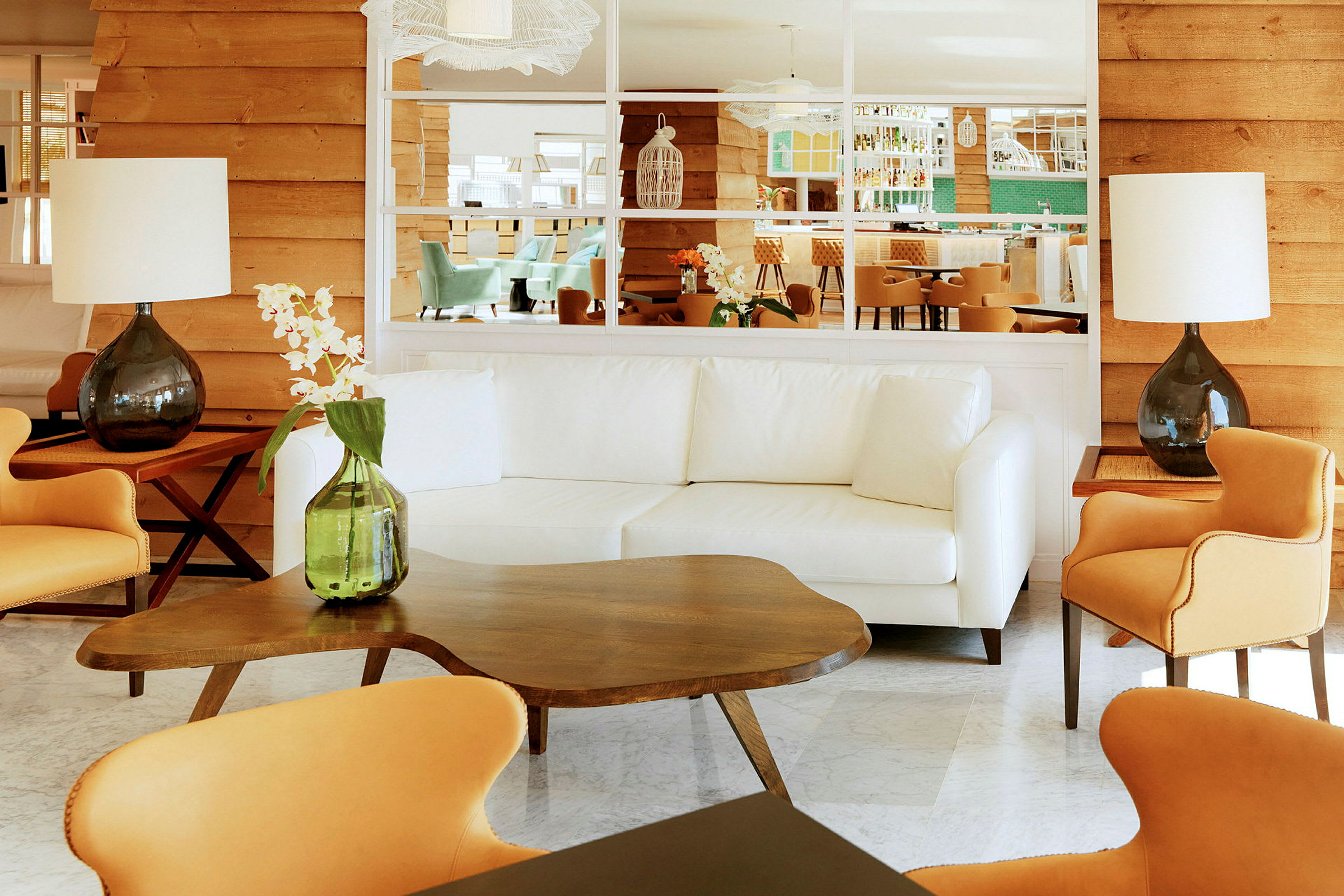 Olohuone, jossa on vaaleita huonekaluja, kuten valkoinen sohva, oransseja tuoleja ja puinen pöytä, sekä koristekasveja ja pöytälamppuja. (Tekoälyn luoma teksti)