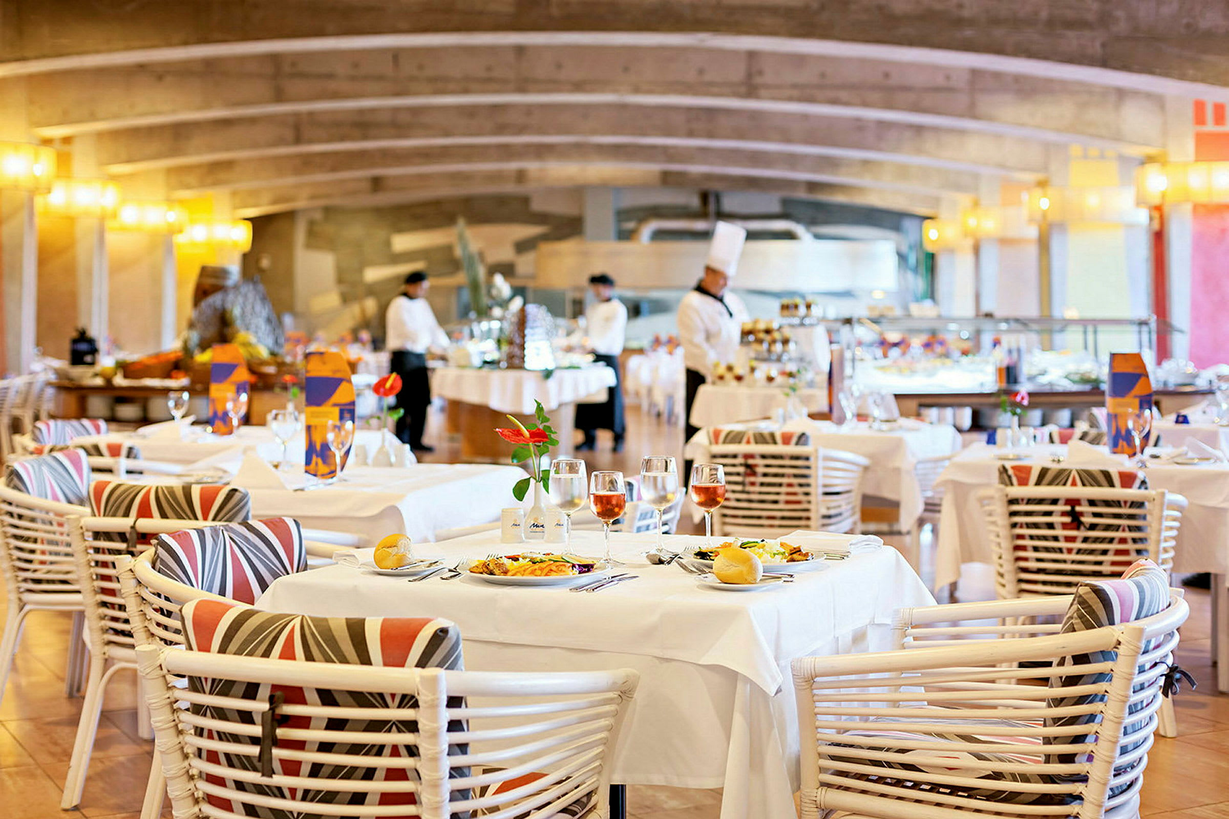 Ravintola-alue, jossa on katettuja pöytiä kukilla ja viinilaseilla koristeltuna. Taustalla henkilökunta työskentelee buffetpöydän luona. (Tekoälyn luoma teksti)