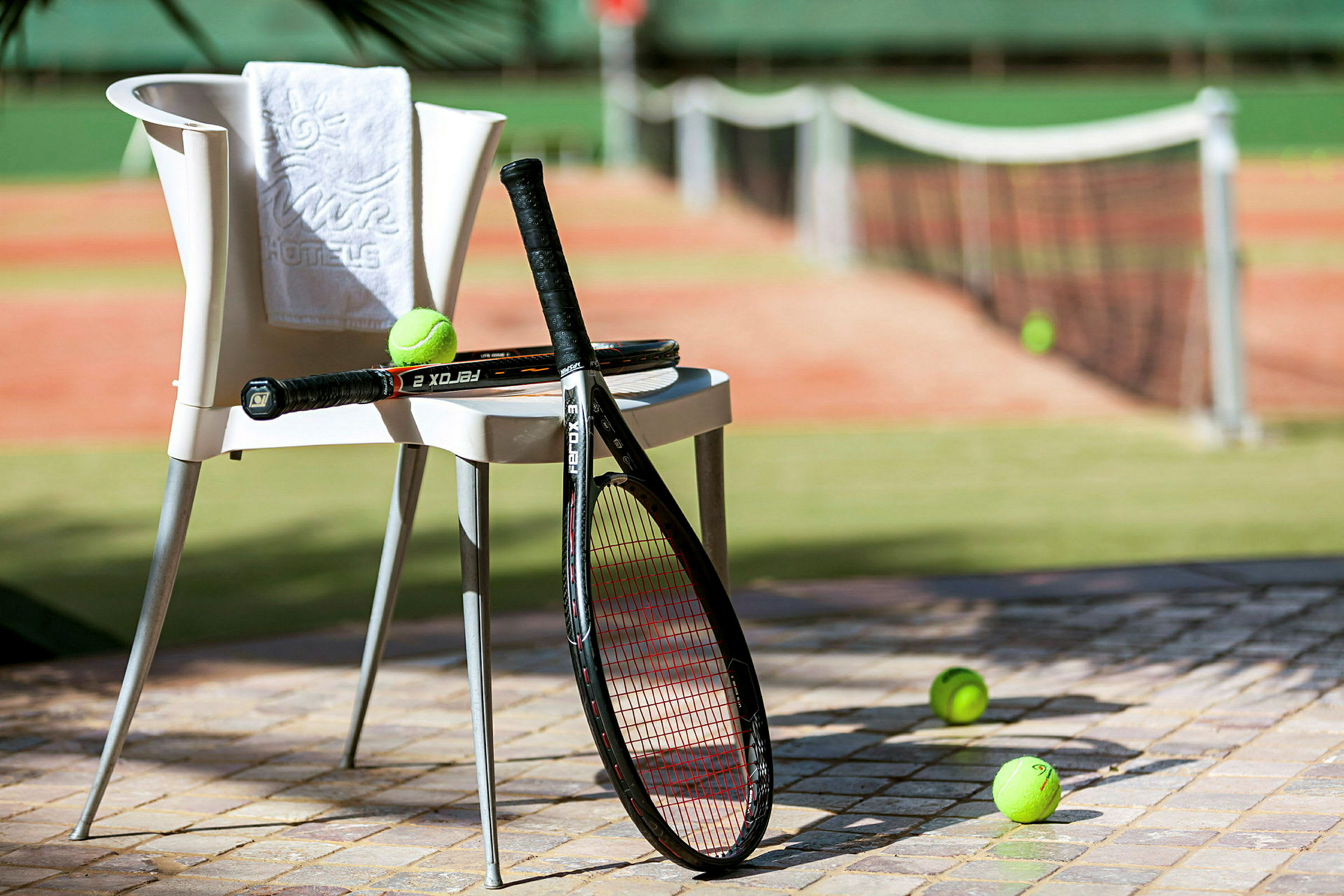 Tuoli, jossa pyyhe ja kaksi tennismailaa, sijaitsee tenniskentän vieressä, ja tennispalloja on maassa. (Tekoälyn luoma teksti)