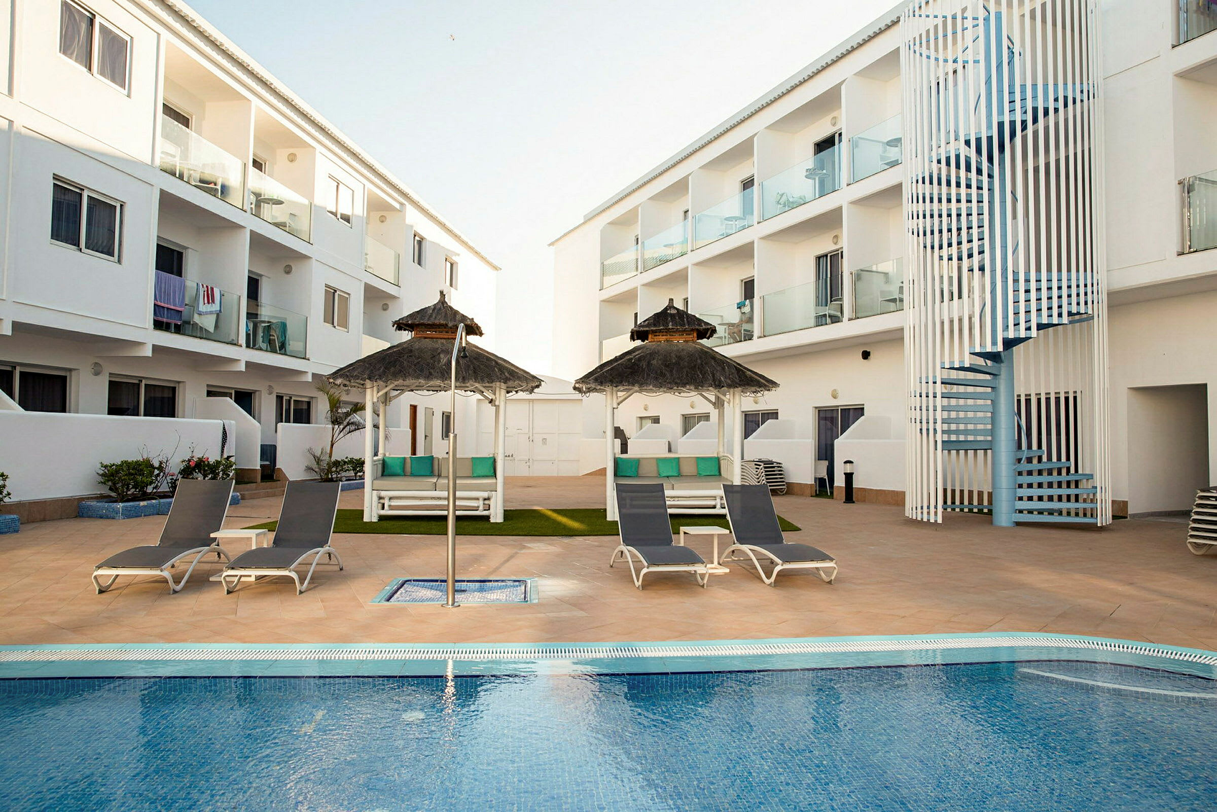 Hotellialue, jossa on uima-allas, ympärillään aurinkotuoleja ja aurinkovarjoja. Taustalla näkyy valkoisia parvekkeellisia rakennuksia. (Tekoälyn luoma teksti)