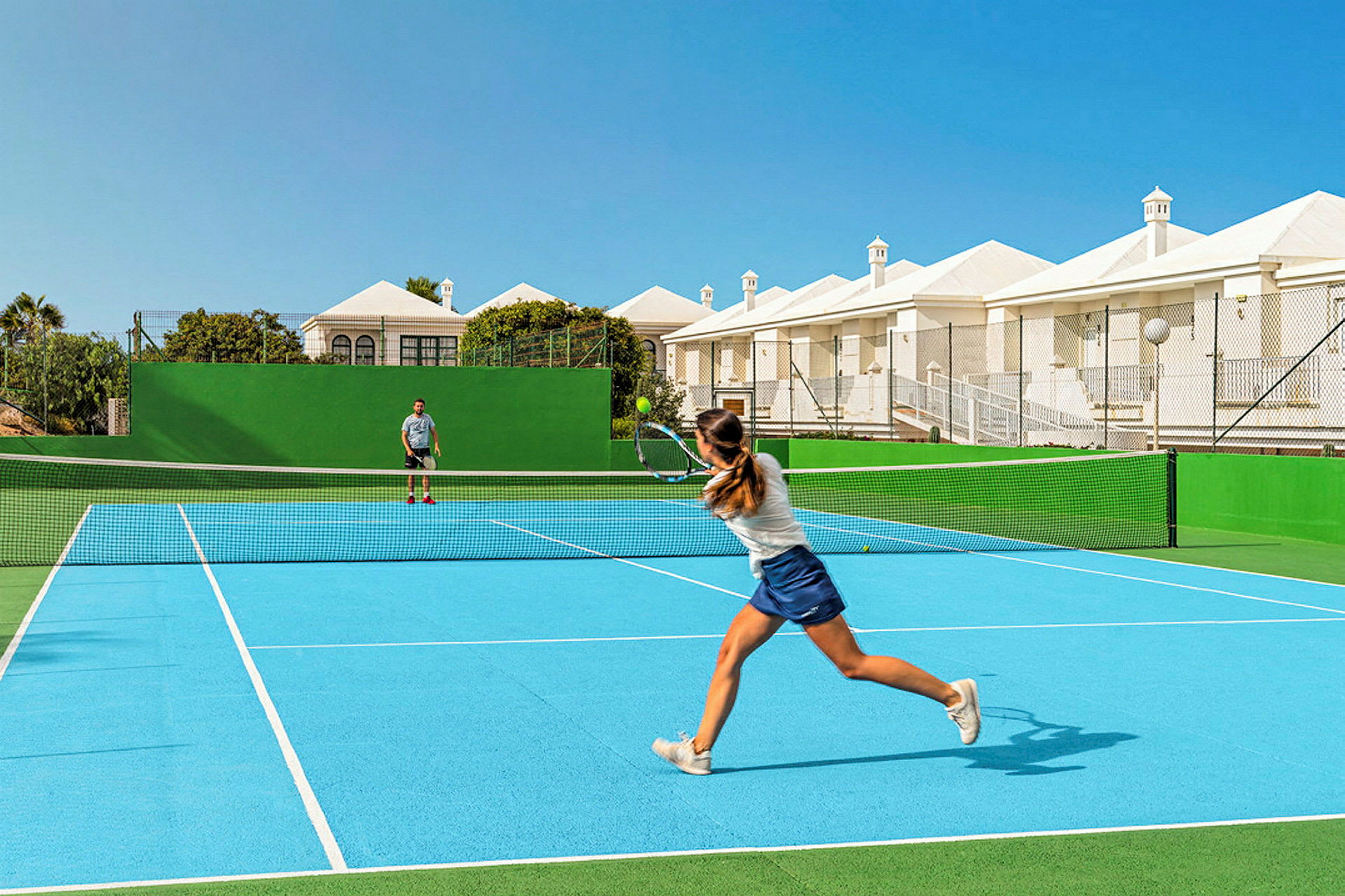Kaksi henkilöä pelaa tennistä ulkokentällä, jolla on sininen alusta ja vihreä ympäristö. Kentän takana näkyy valkoisia rakennuksia ja sininen taivas. (Tekoälyn luoma teksti)