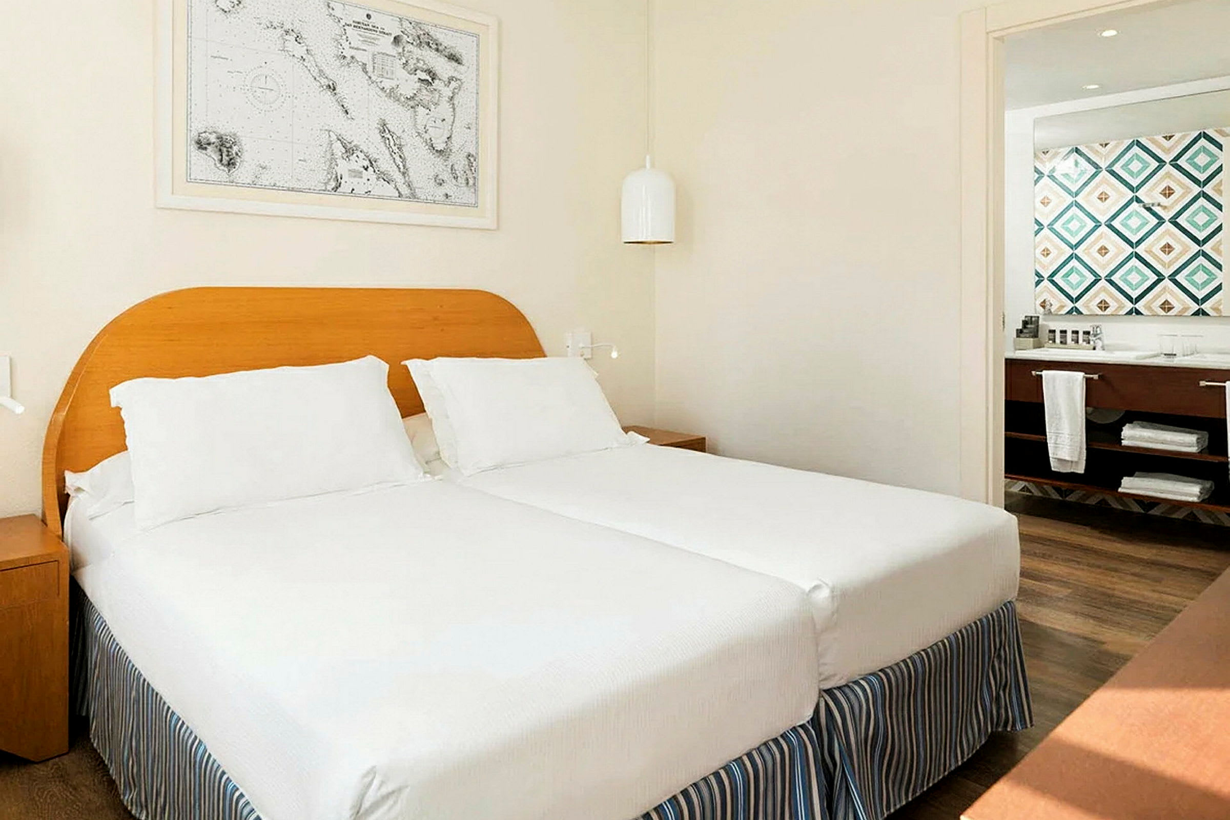 Hotellihuoneen sisustus, jossa kaksi yhteen työnnettyä yhden hengen vuodetta valkoisilla vuodevaatteilla ja vaalea puurunko. Seinällä riippuu taulu. Taustalla näkyy kylpyhuone värikkäine kaakeleineen. (Tekoälyn luoma teksti)