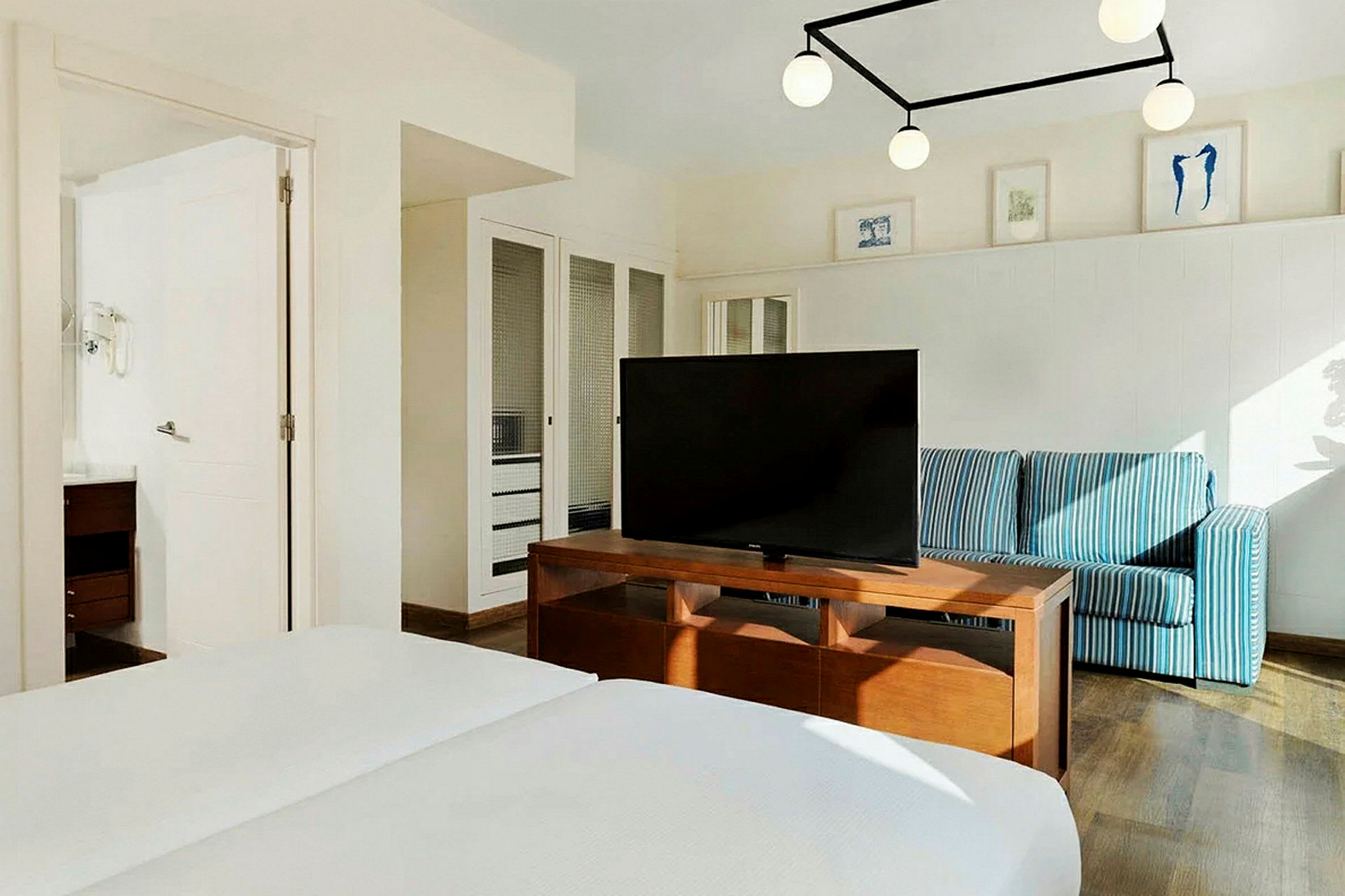 Hotellihuone, jossa on parivuode, televisio puisella pöydällä, sinivalkoraidallinen sohva ja seinäkoristeita. (Tekoälyn luoma teksti)