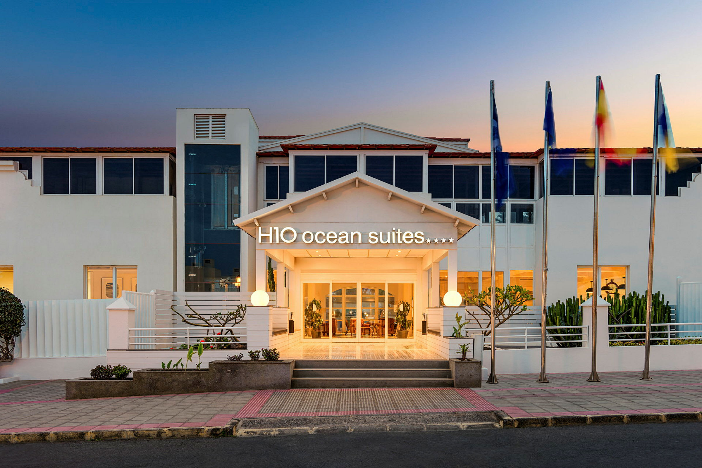 Hotellin sisäänkäynti, jossa H10 Ocean Suites -kyltti ja lipputangot auringonlaskun aikaan. (Tekoälyn luoma teksti)