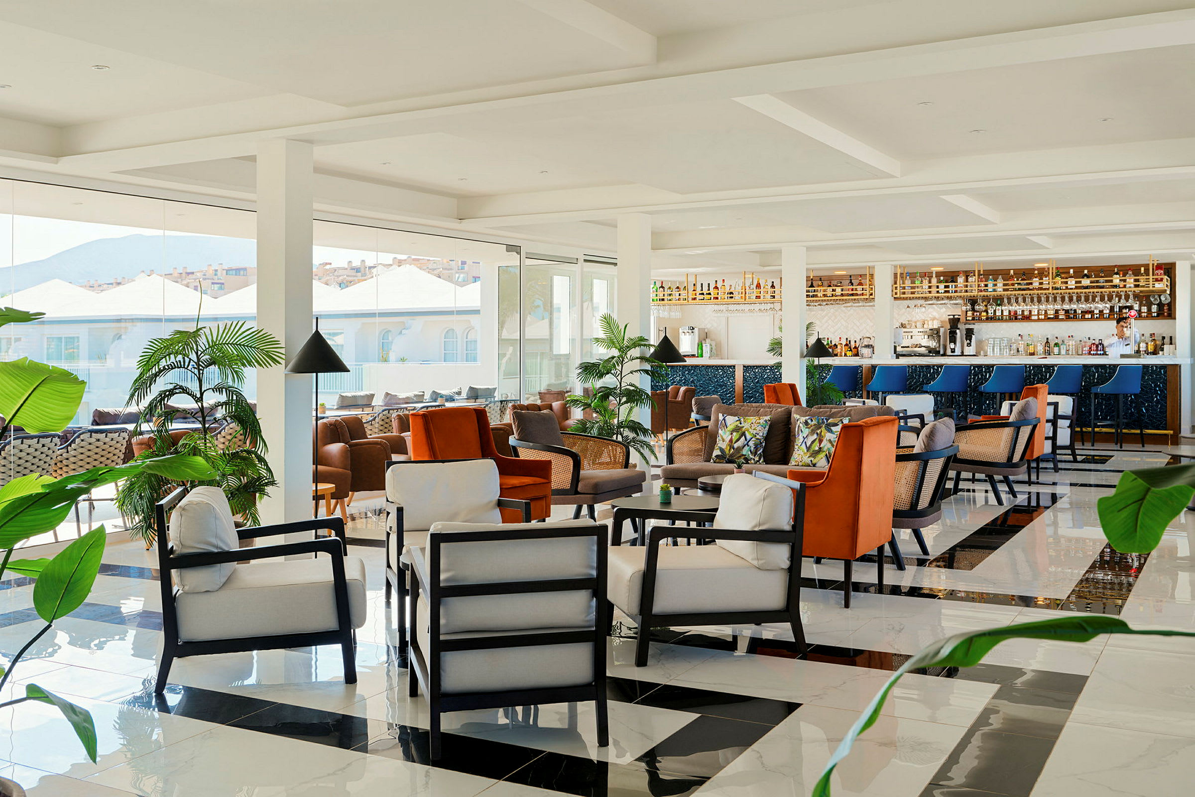 Moderni lounge, jossa mukavia istuimia, baari ja vehreitä kasveja. Suuret ikkunat tarjoavat näkymät rakennuksiin ja vuorille. (Tekoälyn luoma teksti)