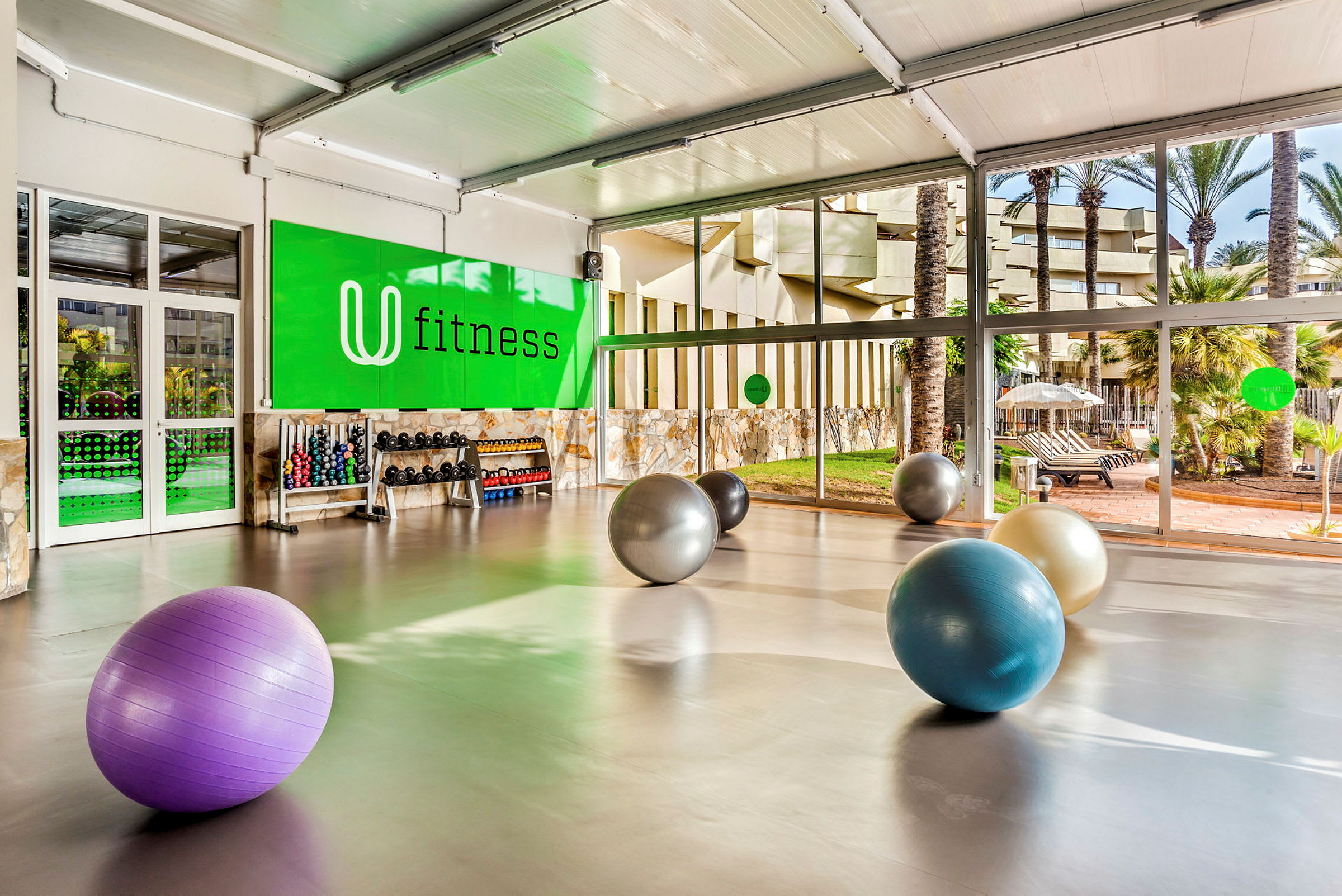 Kuntosali, jossa suuret ikkunat ja kuntopalloja lattialla. Käsipainotelineitä ja kuntoiluvälineitä näkyy vihreää seinää vasten, jossa lukee "fitness". (Tekoälyn luoma teksti)
