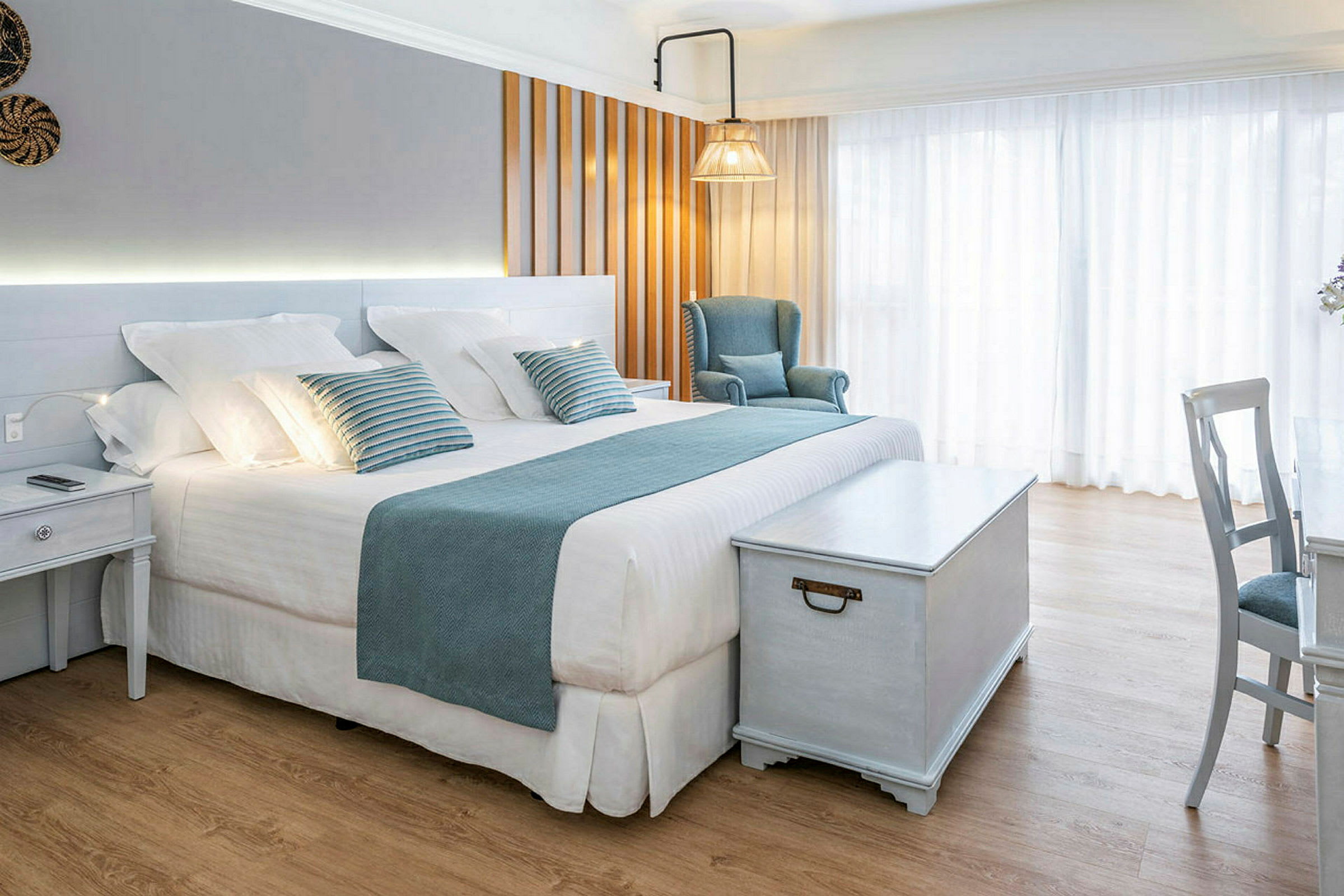 Vaalea ja tyylikäs hotellihuone, jossa on iso sänky, sinisiä koristeita, nojatuoli ja puupöytä. (Tekoälyn luoma teksti)