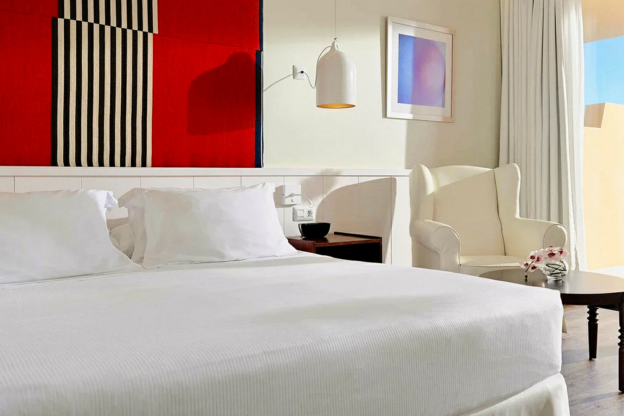 Tyylikäs hotellihuone, jossa on iso sänky ja valkoiset vuodevaatteet, nojatuoli pienen pöydän vieressä sekä värikäs seinäkoriste sängyn yläpuolella. (Tekoälyn luoma teksti)